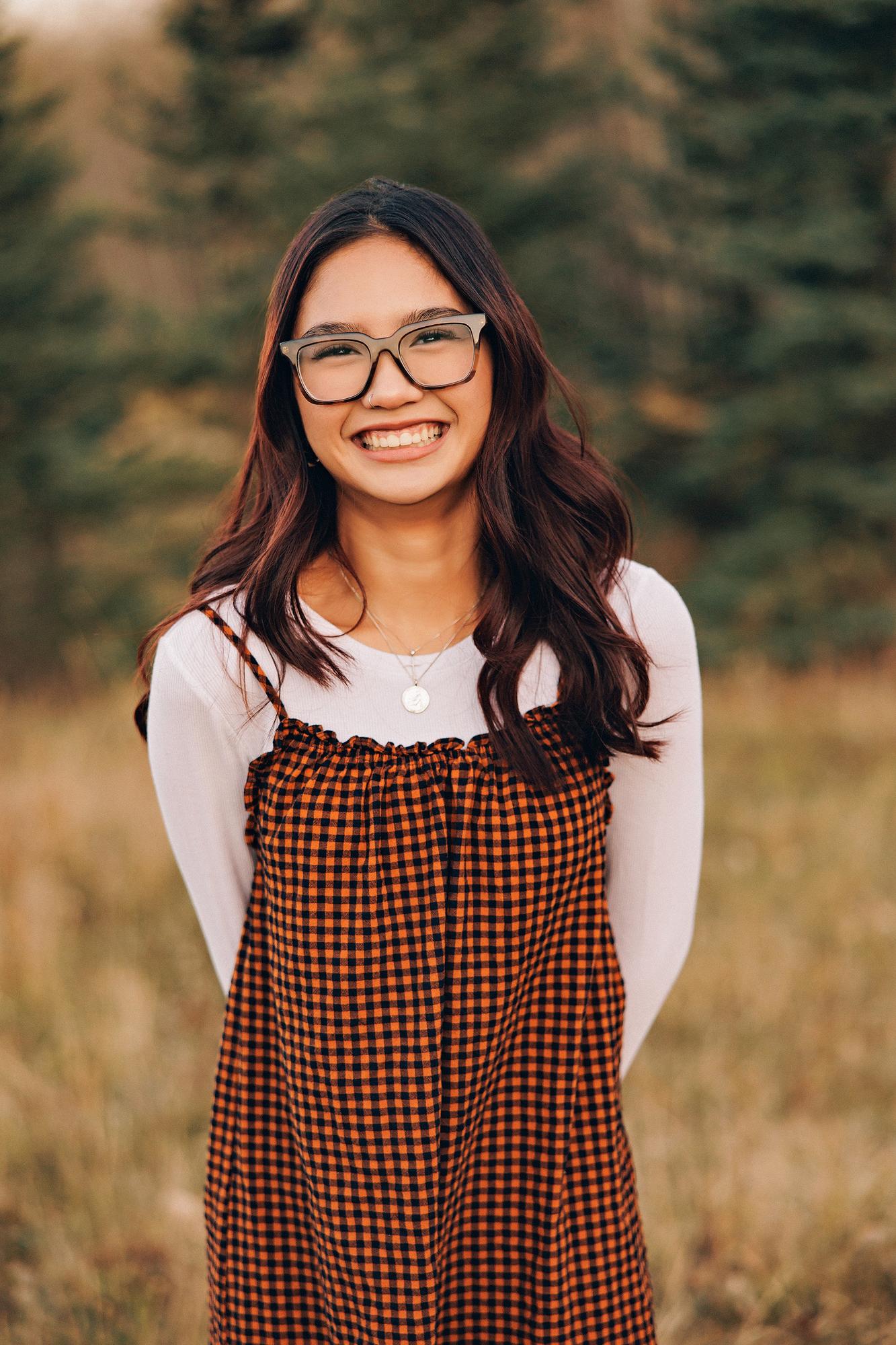 Outdoor family photography Calgary fall: Close-up portrait of a teenage girl with dark wavy hair and bold black-frame glasses wearing an orange gingham ruffle sundress over a white long-sleeve top with a delicate gold necklace, smiling brightly in front of tall evergreen trees