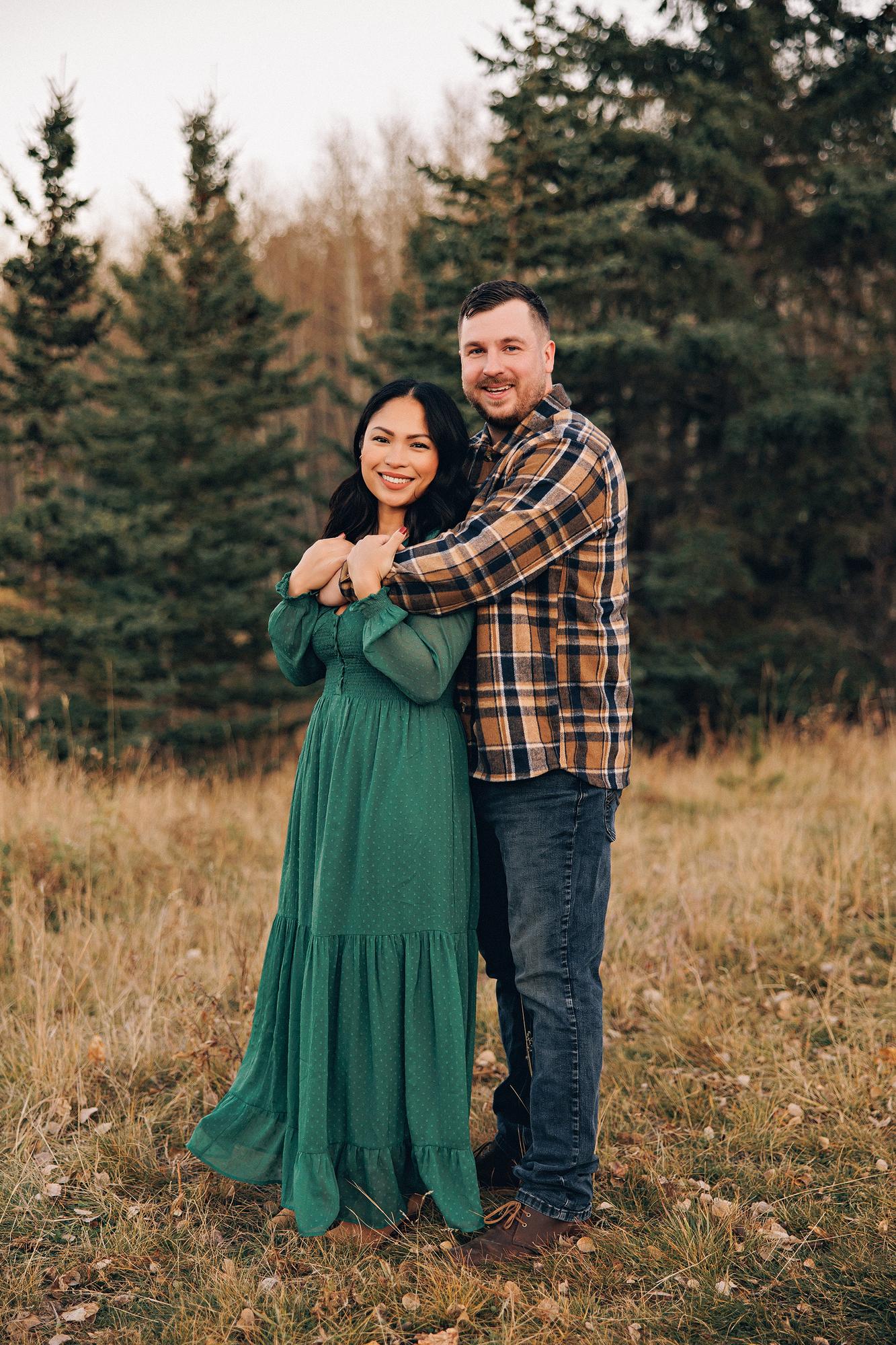 Outdoor couple photography Calgary fall: Smiling couple embracing and facing camera in a golden meadow — woman in an emerald green maxi dress and man in a brown plaid flannel shirt with his arms wrapped around her, tall evergreen trees behind them