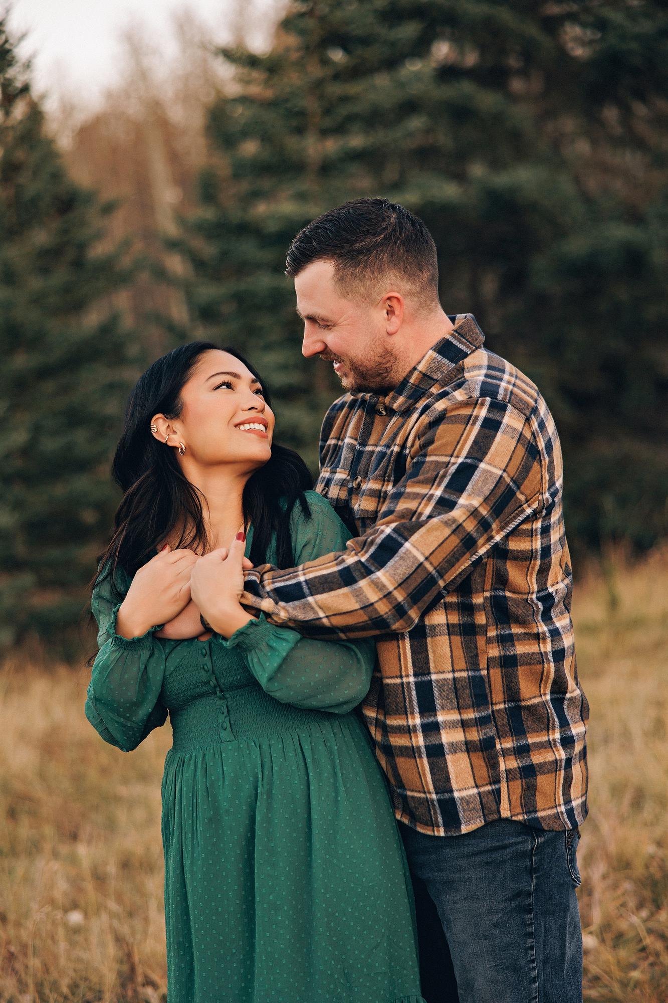 Outdoor couple photography Calgary fall: Romantic close-up of couple gazing at each other and laughing — woman in an emerald green dress looking up with a radiant smile, man in brown plaid flannel smiling down at her, evergreen trees in warm golden light behind them