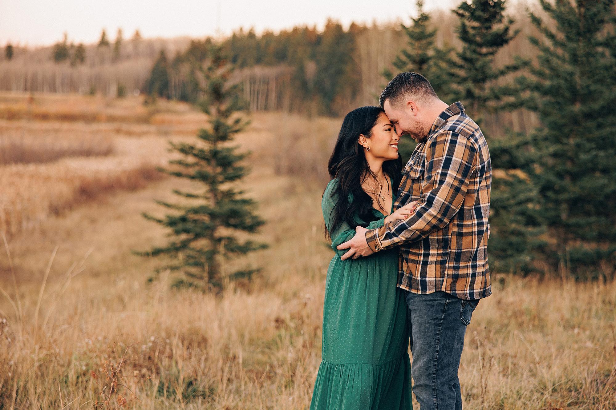 Outdoor couple photography Calgary fall: Intimate full-length portrait of couple touching foreheads and embracing in a wide open golden meadow — woman in emerald green maxi dress, man in brown plaid flannel — small evergreen trees and autumn forest stretching behind them