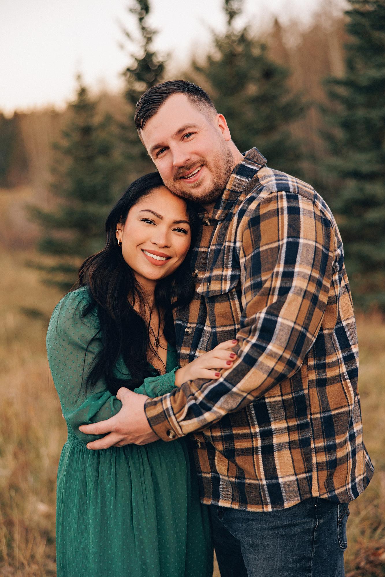 Outdoor couple photography Calgary fall: Warm close-up of couple cheek-to-cheek smiling at camera — woman in an emerald green dress with long dark wavy hair, man in brown plaid flannel — soft bokeh evergreen trees behind them