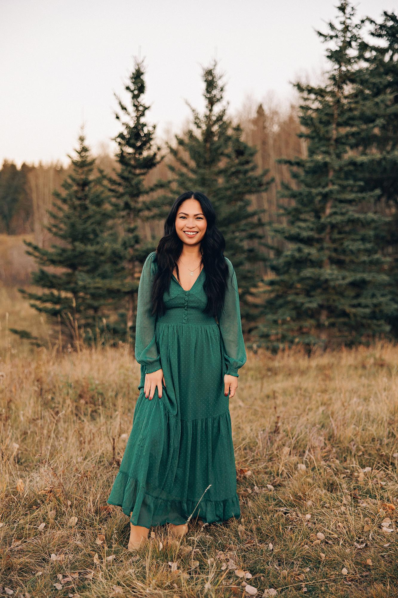 Outdoor family photography Calgary fall: Full-length portrait of a smiling woman with long dark wavy hair in an emerald green long-sleeve maxi dress with a gold necklace, standing confidently in a golden meadow with tall evergreen trees behind her