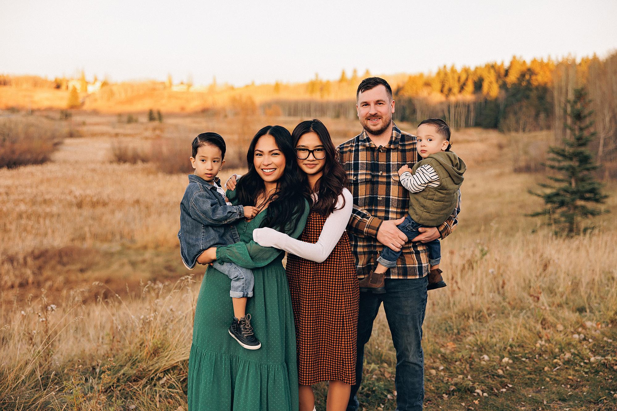 Outdoor family photography Calgary fall: Warm golden hour portrait of a family of five in a sunlit open field — mother in emerald green dress holding toddler son, father in plaid flannel holding baby, teenage daughter in orange gingham between them — evergreen and autumn trees behind them