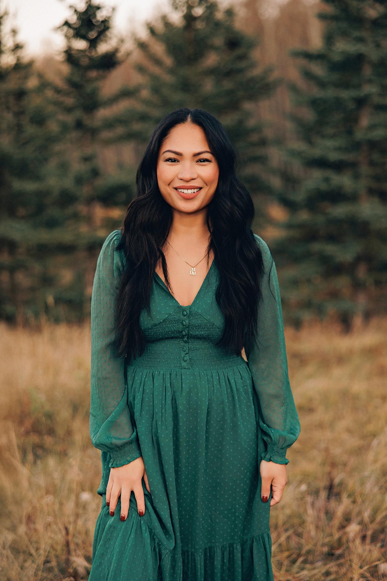 Outdoor family photography Calgary fall: Close-up portrait of a smiling woman with long dark wavy hair in an emerald green V-neck maxi dress and gold name necklace, evergreen trees softly bokeh behind her in warm autumn light