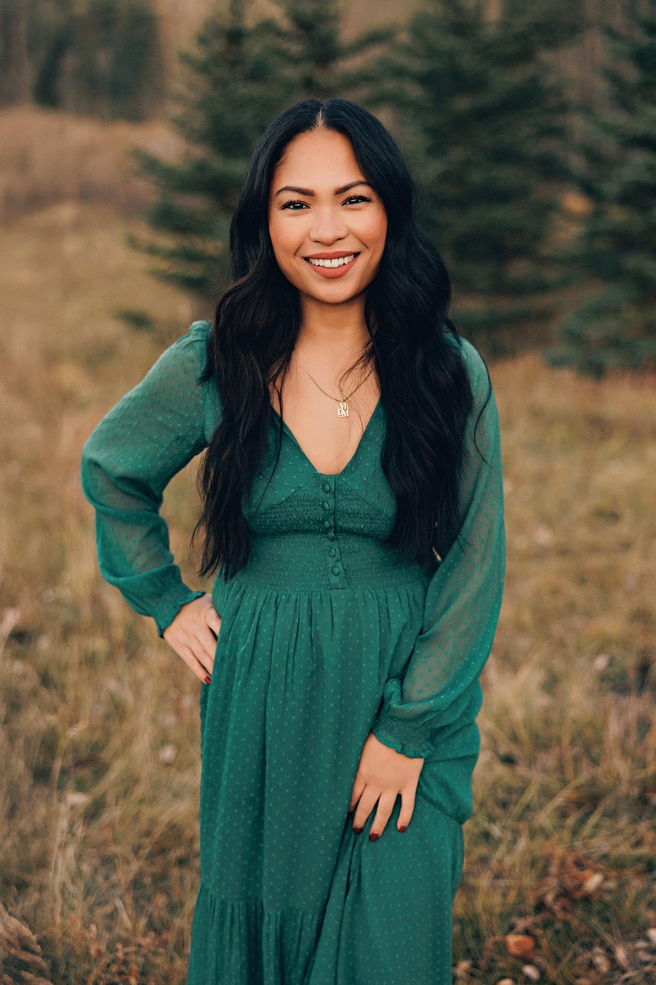 Outdoor family photography Calgary fall: Close-up portrait of a radiant woman with long dark wavy hair in an emerald green V-neck maxi dress and gold name necklace, hand on hip and a bright smile, evergreen trees behind her