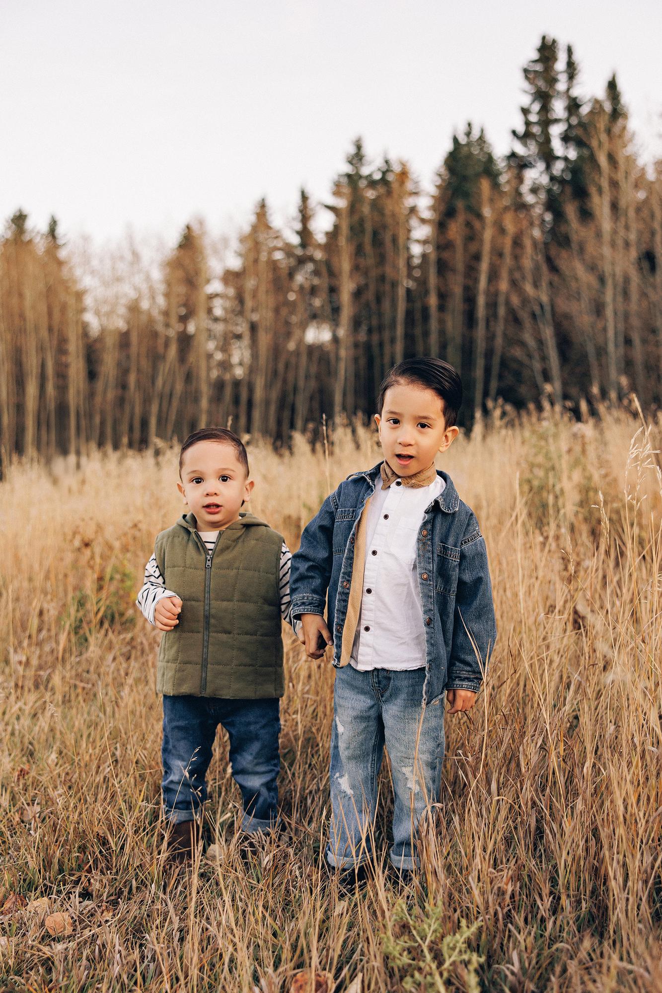 Outdoor family photography Calgary fall: Two young brothers standing hand-in-hand in golden meadow grass — toddler in an olive puffer vest and striped top, older boy in a denim jacket with bow tie — autumn birch forest behind them