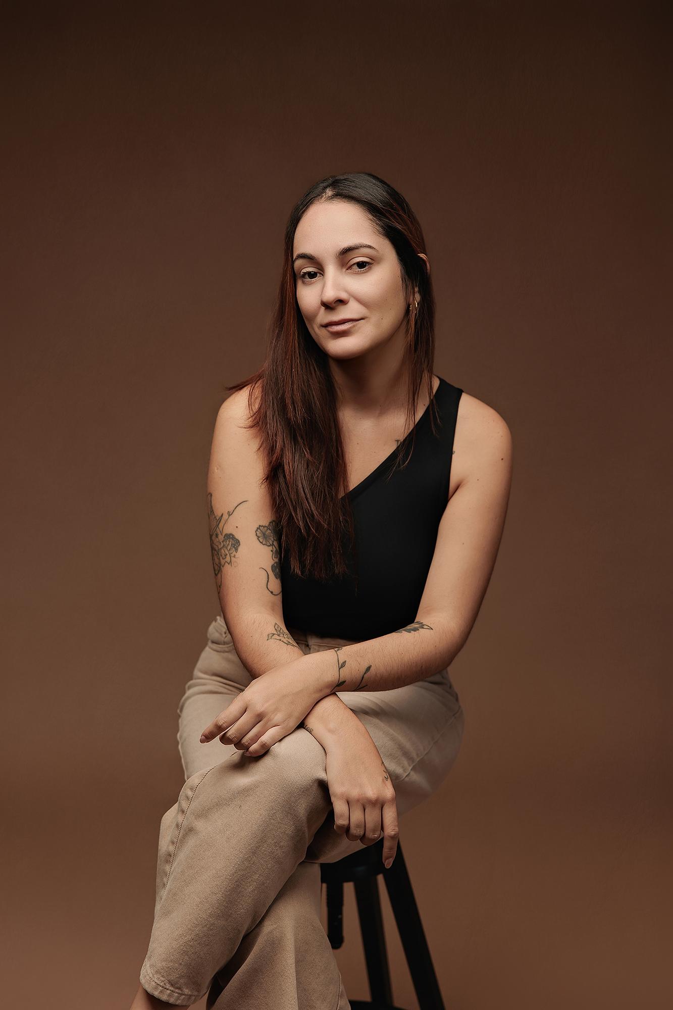 A woman with long dark hair and tattoos poses confidently in a Calgary studio, wearing a black top against a warm, professional brown backdrop.