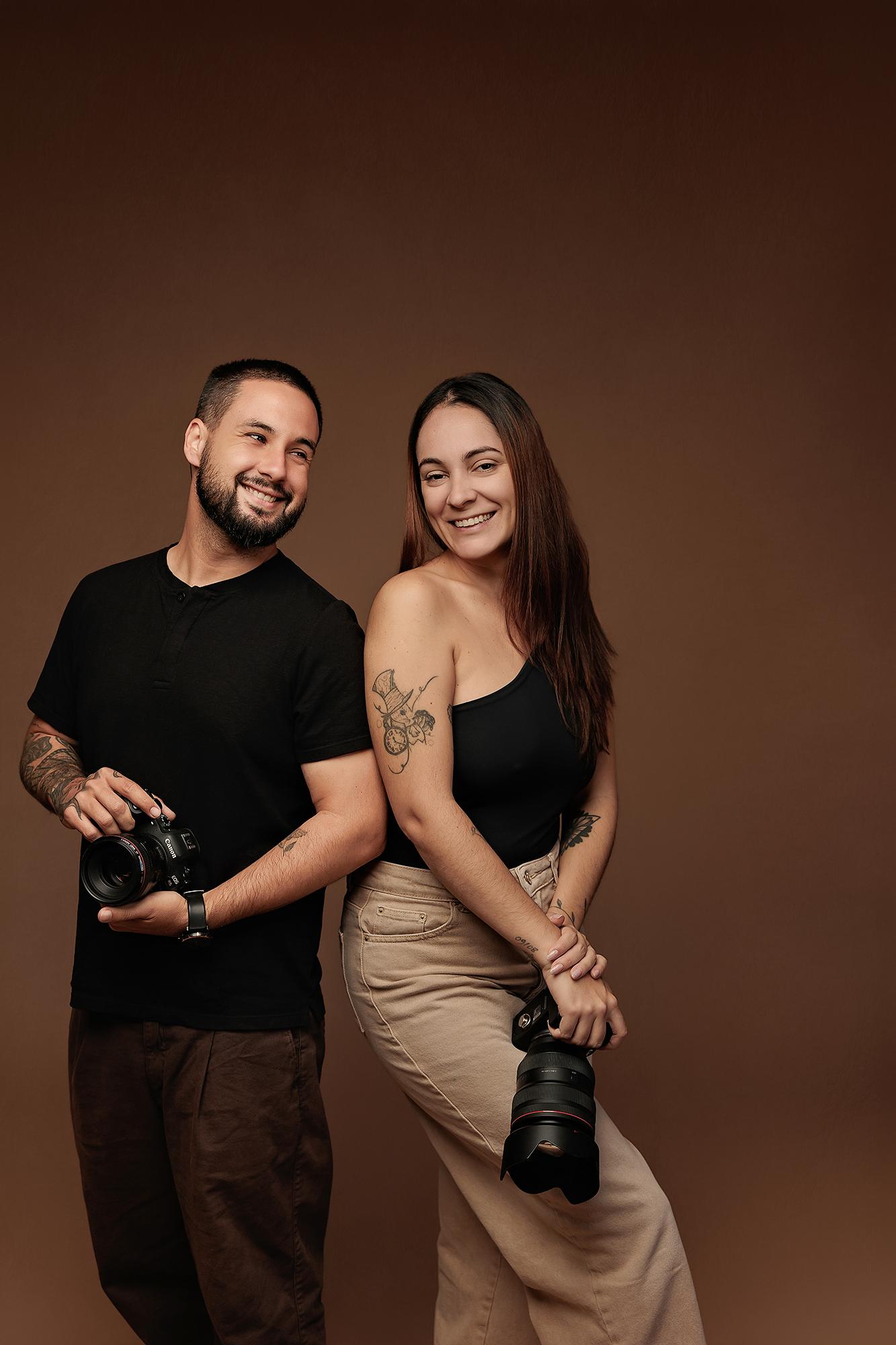 A smiling man and woman stand back-to-back holding cameras in this Calgary photography studio, showcasing the professional team behind our headshots.