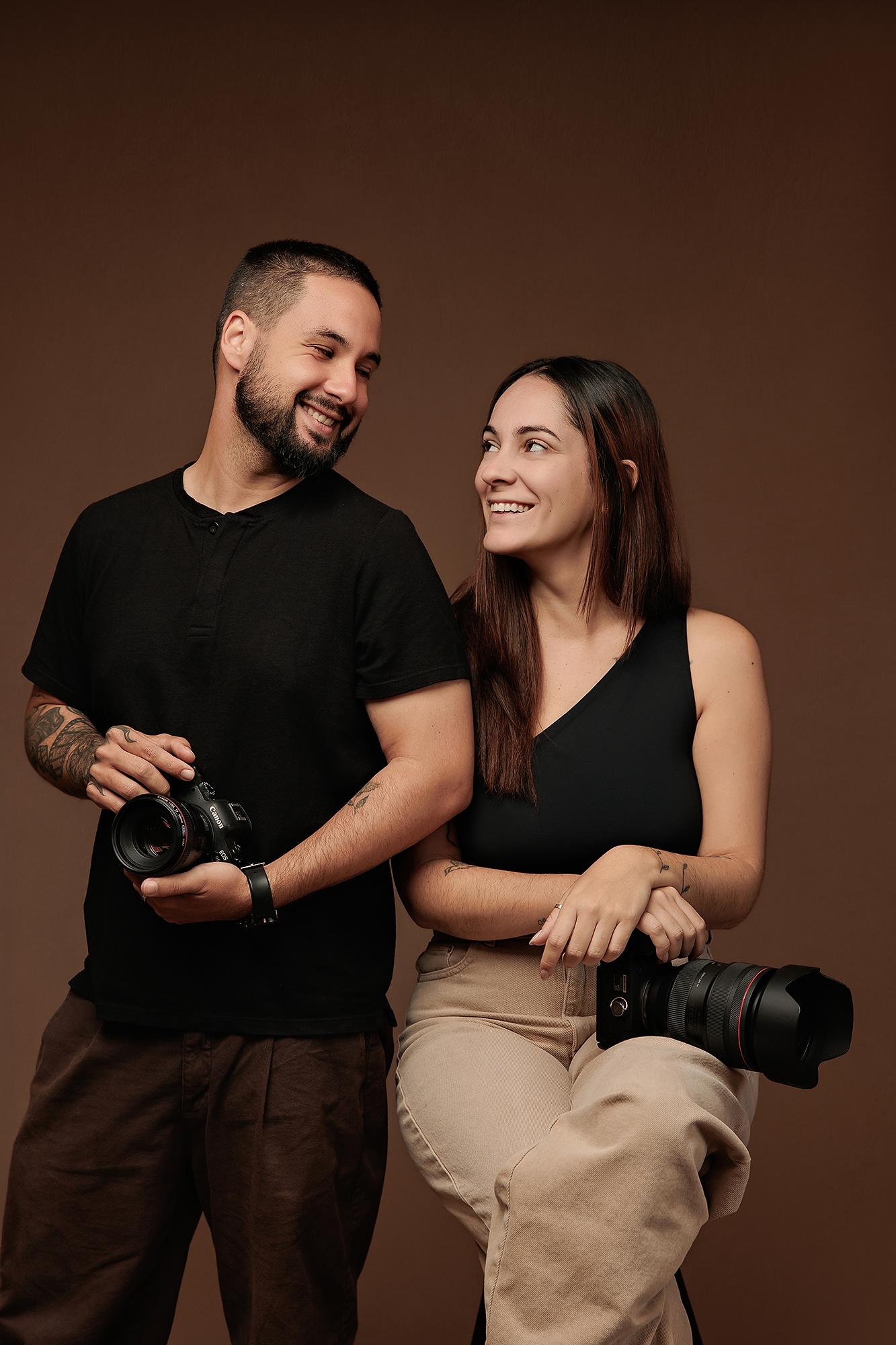 A smiling man and woman hold cameras in this Calgary studio portrait, showcasing the collaborative team behind our professional photography services.