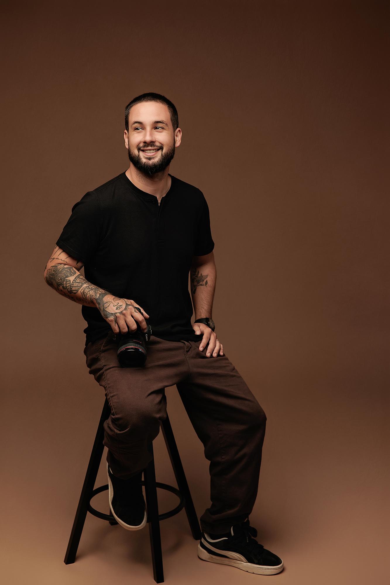 A bearded photographer with arm tattoos smiles while holding a camera in this Calgary studio portrait, set against a warm brown backdrop.