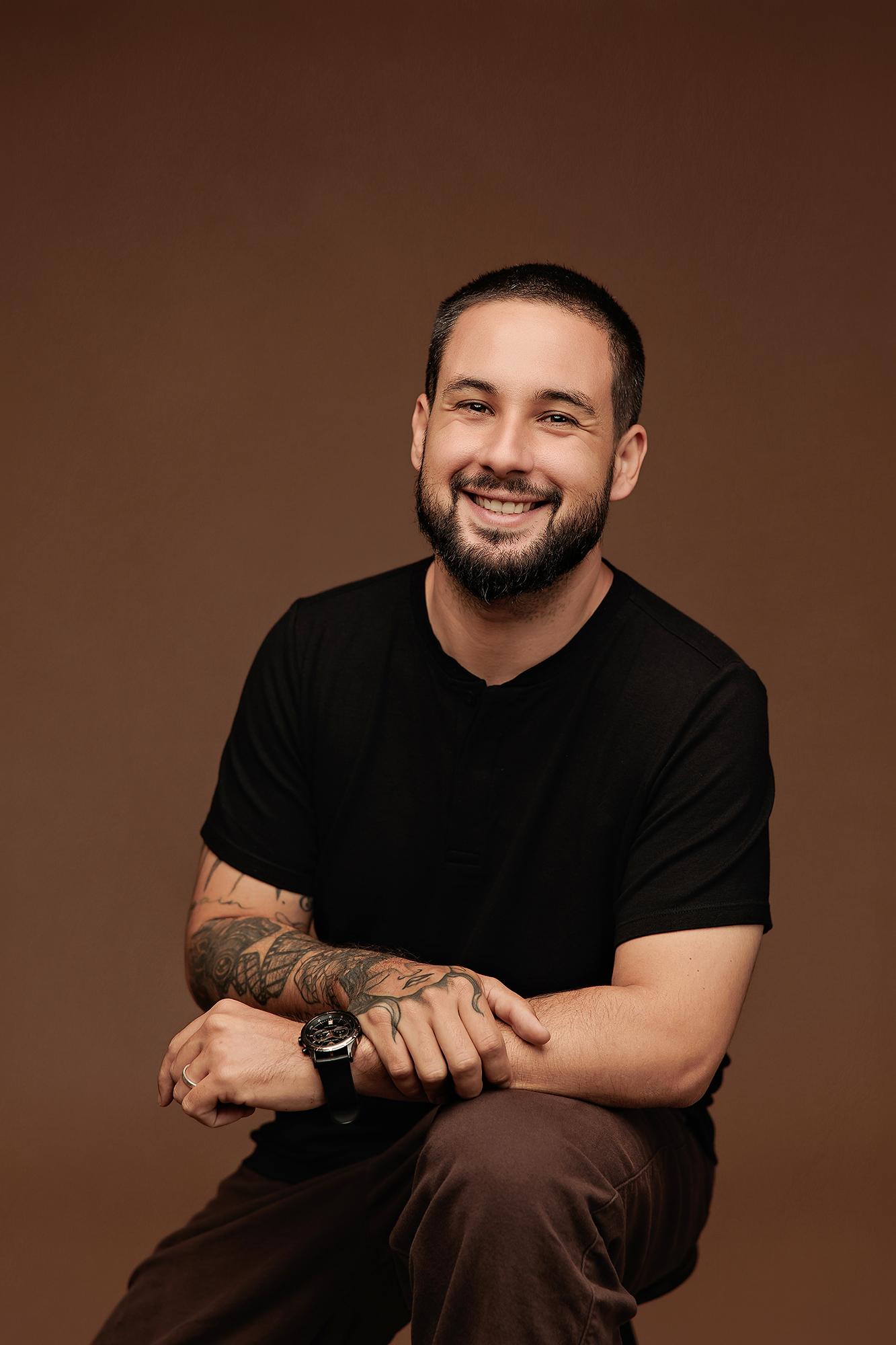 A smiling man with tattoos and a black t-shirt poses for a professional headshot at our Calgary photography studio, featuring soft studio lighting.