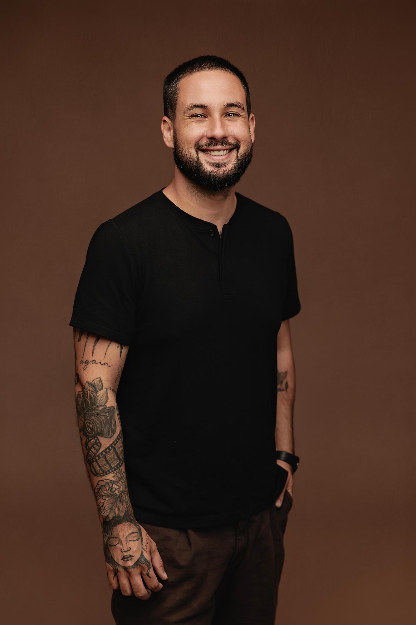 A smiling man with a beard and tattoos wears a black shirt in this professional headshot from a Calgary photography studio, featuring soft studio lighting.