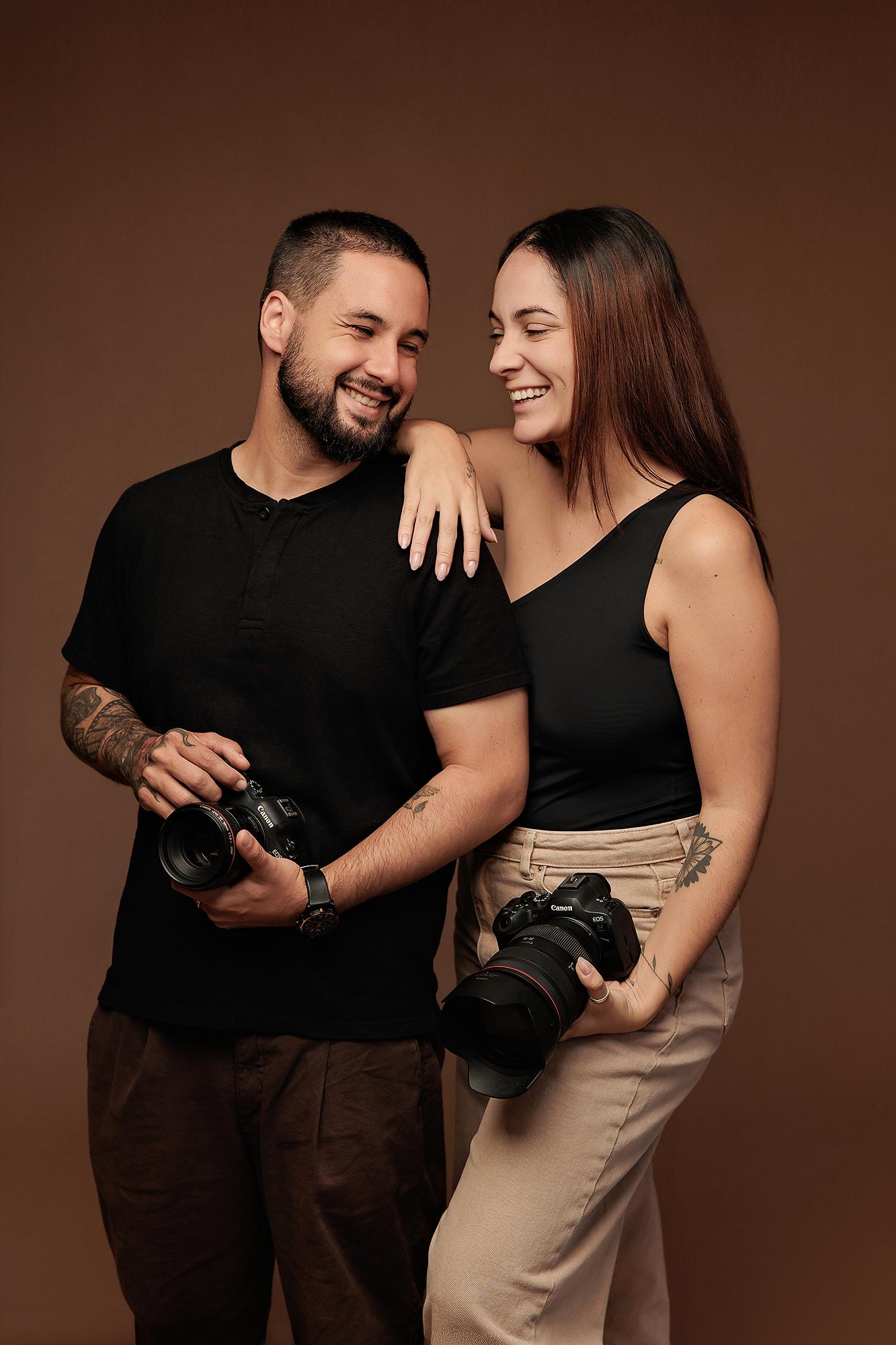 A smiling man and woman hold professional cameras in this Calgary photography studio, showcasing the warm, collaborative energy of our team.