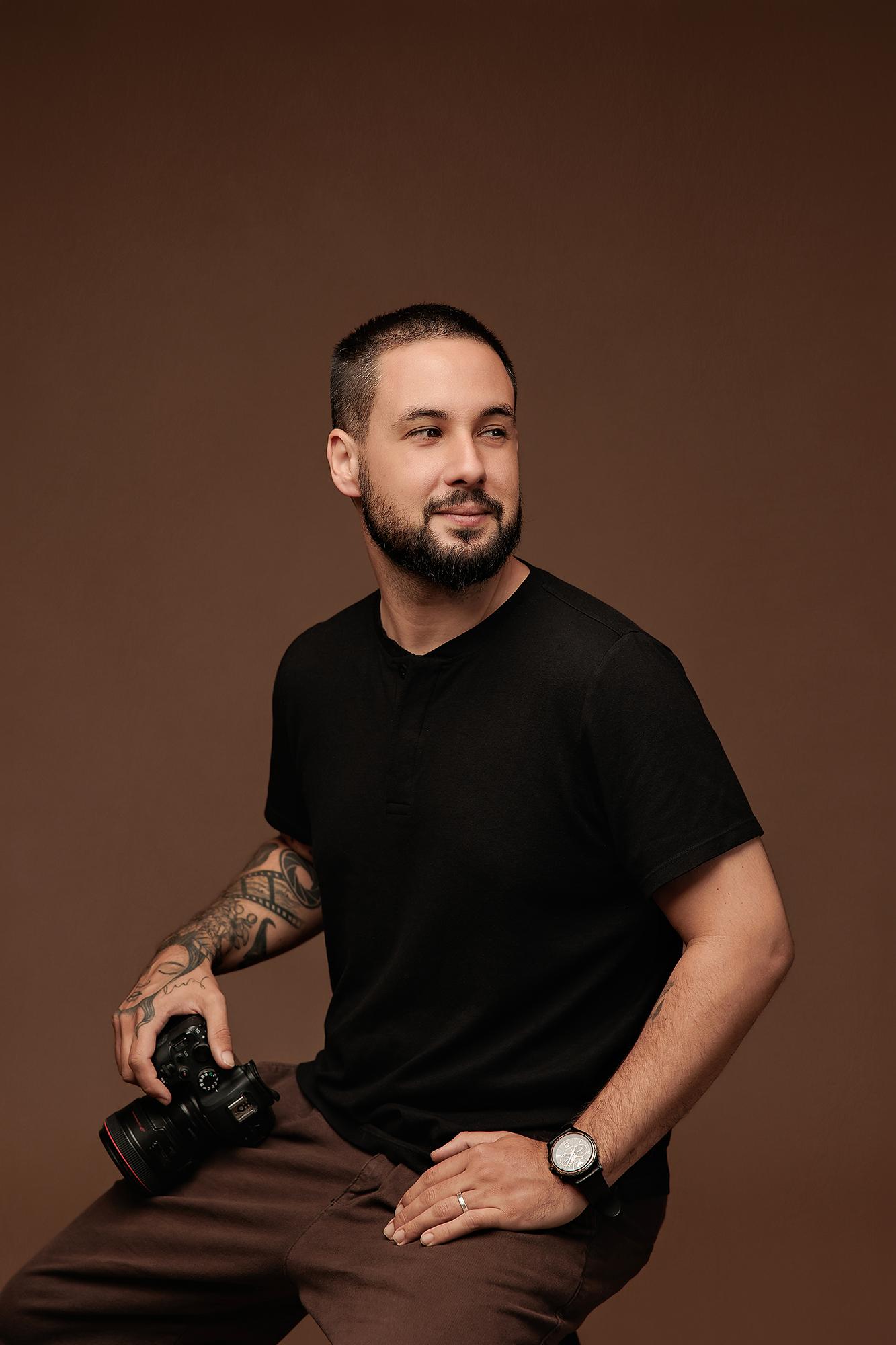A bearded photographer with tattooed arms holds a professional camera in this Calgary studio portrait, featuring soft lighting against a warm brown backdrop.