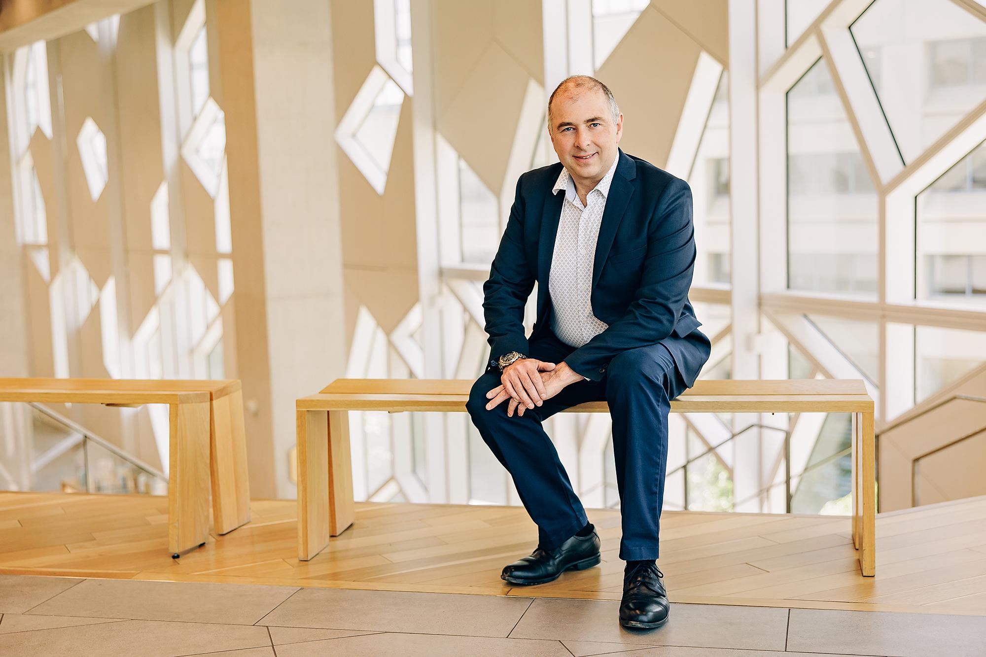 A smiling professional man in a navy suit sits on a wooden bench in a modern Calgary studio, showcasing the clean lighting of our corporate headshots.