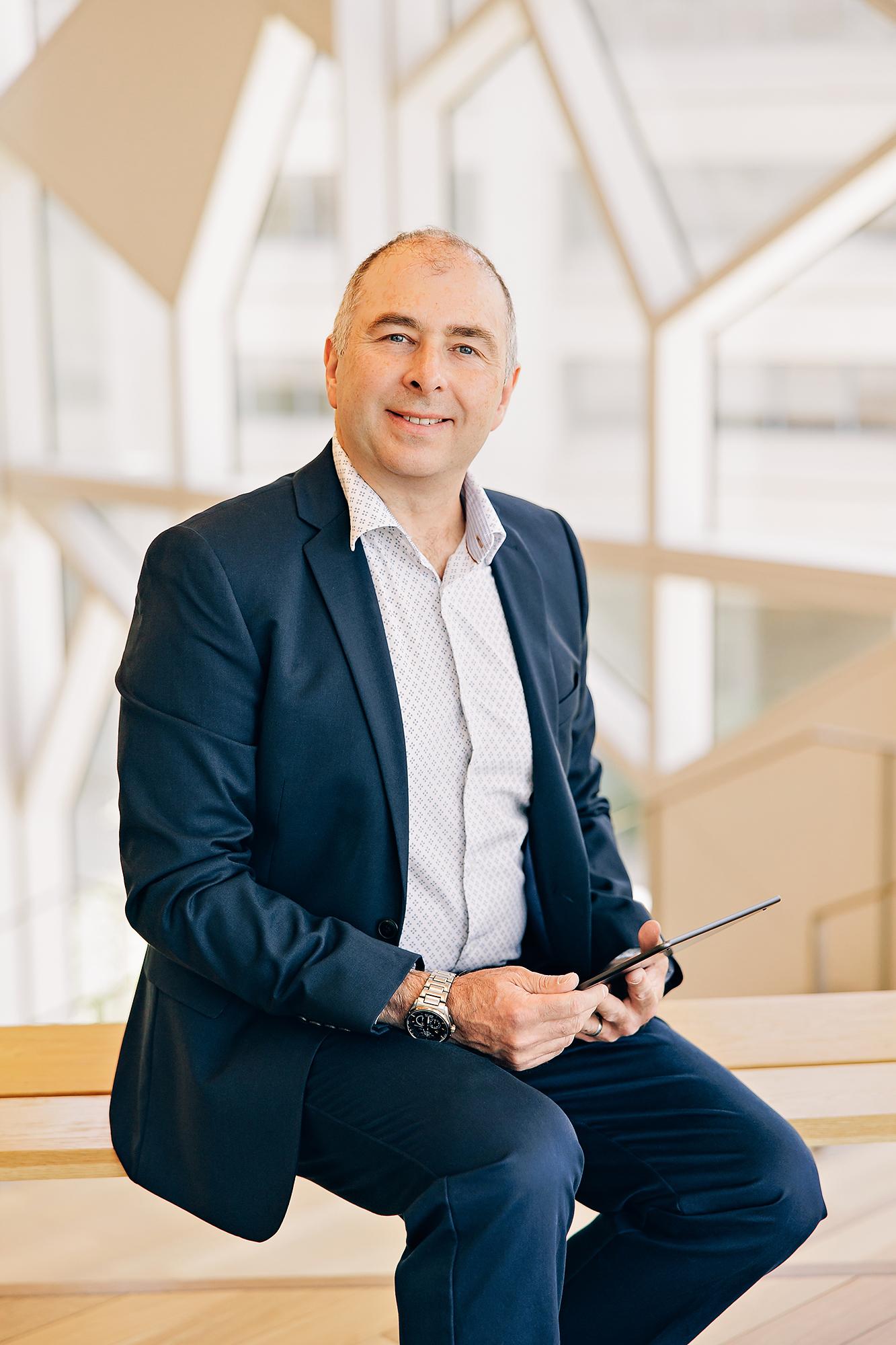 A smiling businessman in a navy suit and patterned shirt sits with a tablet in a bright Calgary studio, showcasing professional corporate branding.