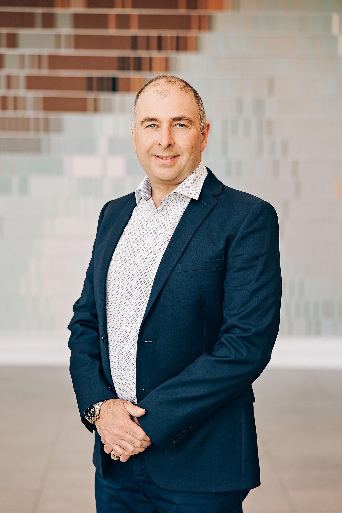 A smiling man in a navy blazer and patterned shirt stands for a professional headshot captured by our Calgary photography studio in soft, bright light.