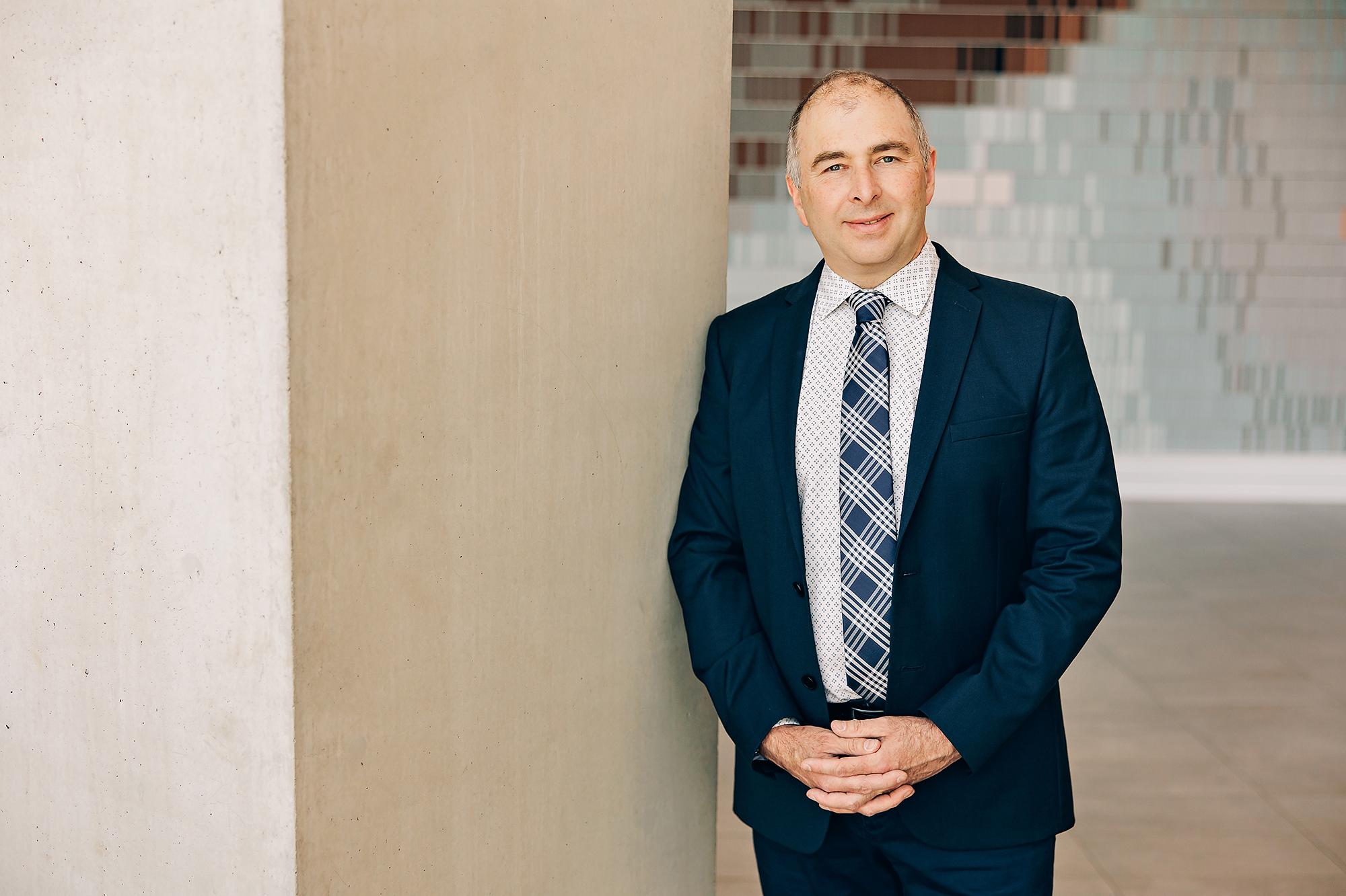 A professional man in a navy suit leans against a concrete pillar, showcasing the modern corporate style of this Calgary headshot photography studio.