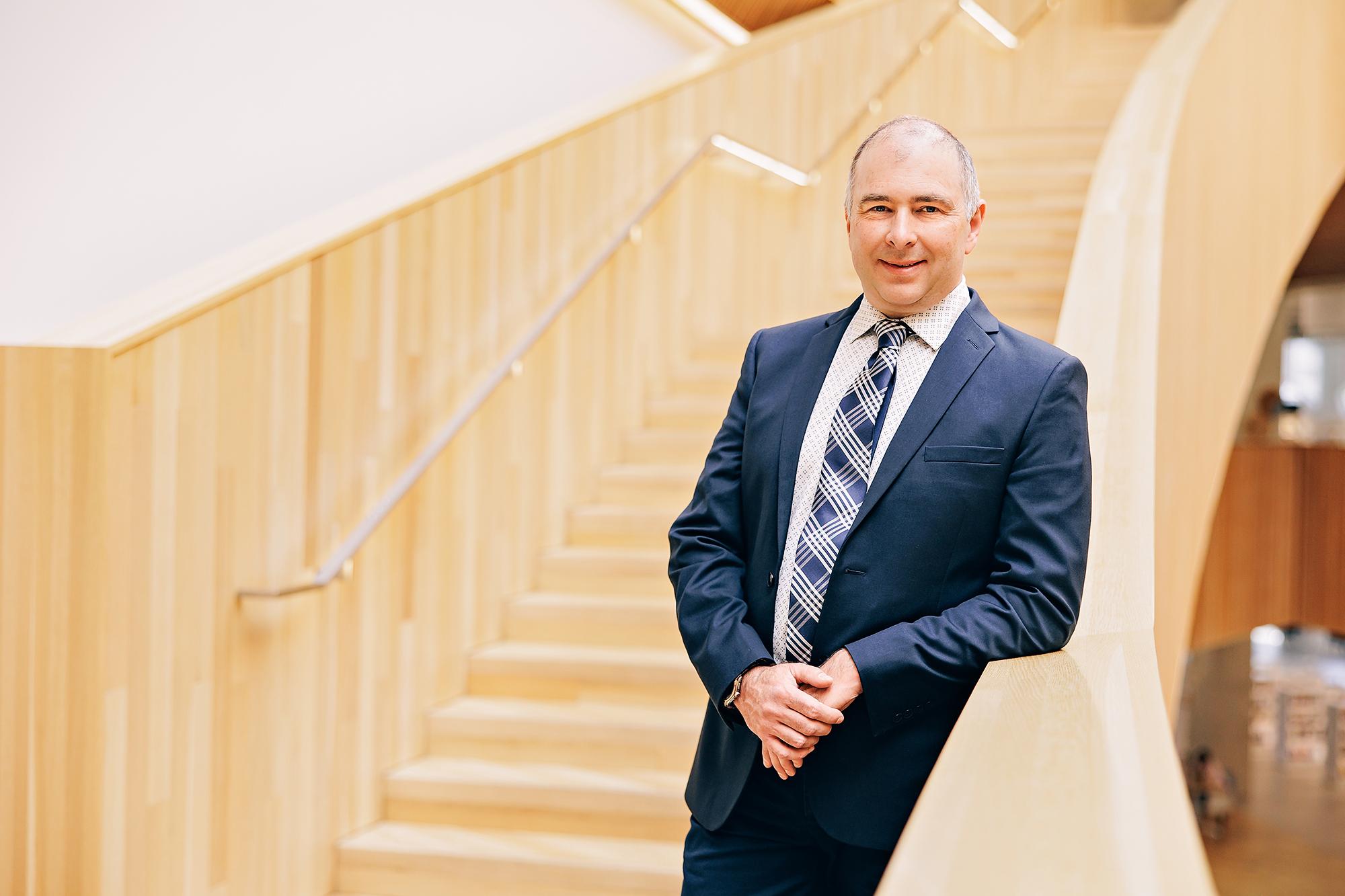 A smiling professional in a navy suit leans against a wooden railing, showcasing the expert corporate headshot services of our Calgary photography studio.