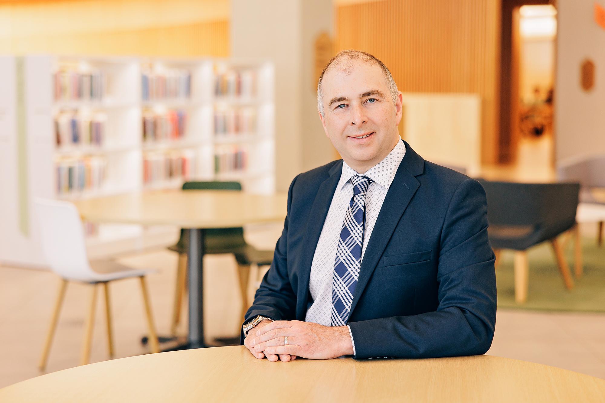 A middle-aged man in a navy suit smiles at a library table, captured by a Calgary professional headshot photographer for a polished corporate look.