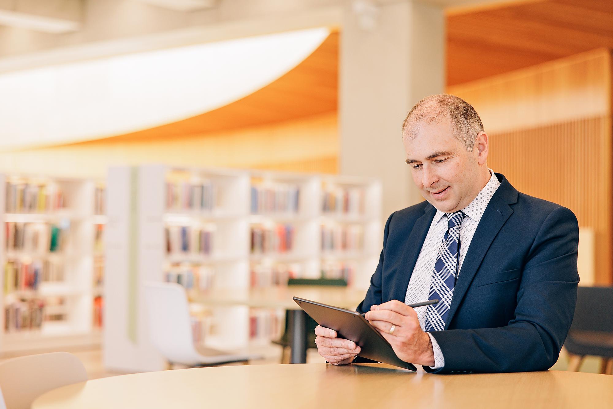 A smiling professional in a navy suit uses a tablet at a library, showcasing the quality of our Calgary headshot photography for personal branding.