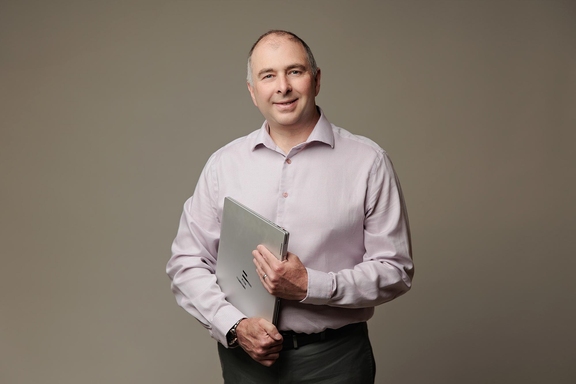 A smiling man in a patterned dress shirt holds a laptop in this professional headshot captured at our Calgary photography studio with soft lighting.