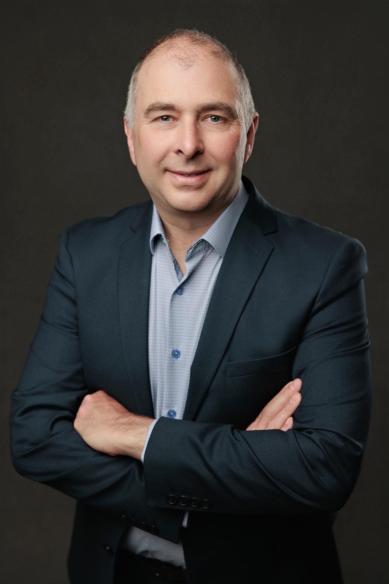 A professional man in a navy blazer smiles with arms crossed, showcasing the polished style of our Calgary headshot photography studio.