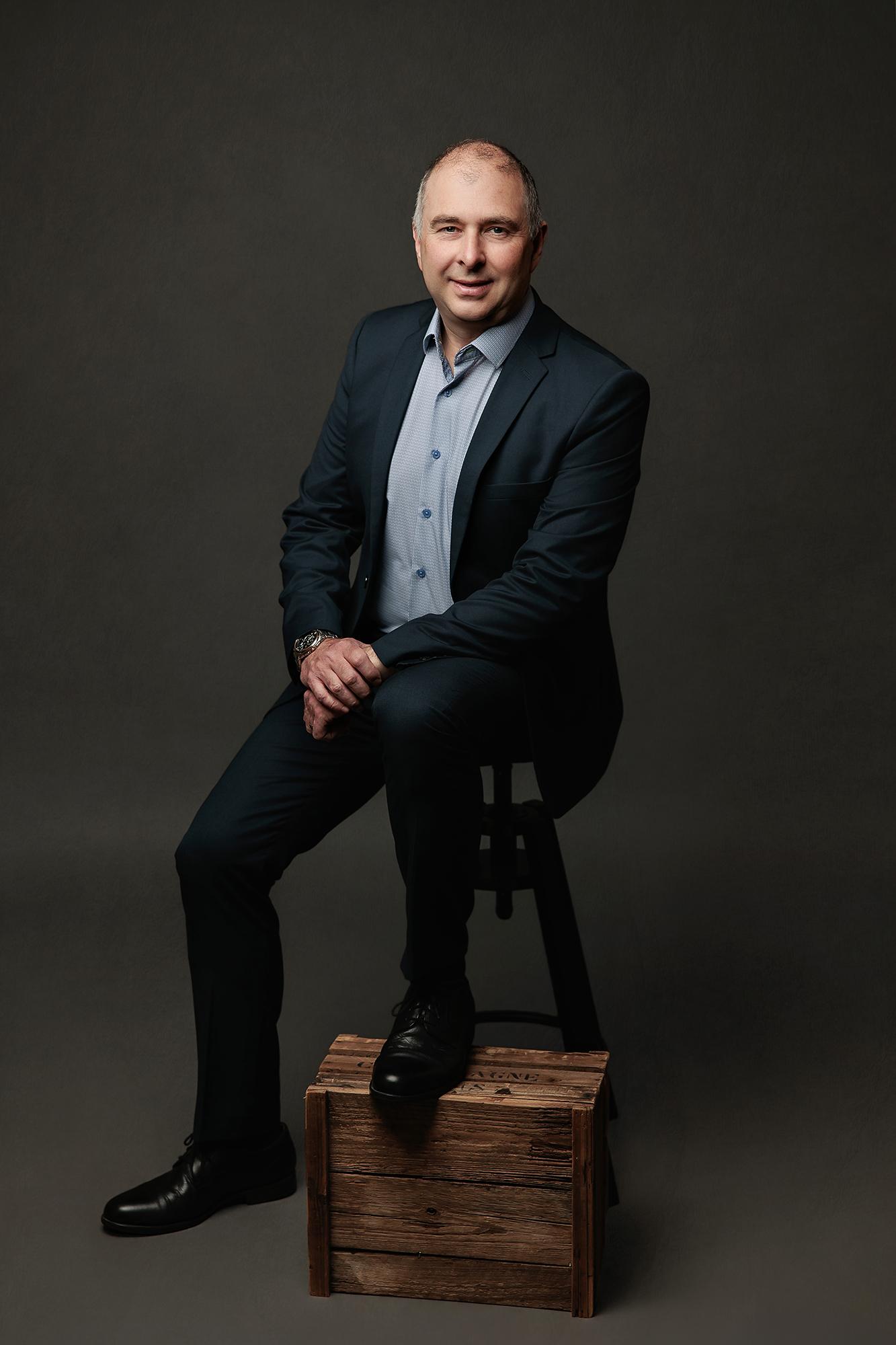 A professional man in a navy suit sits on a wooden crate in our Calgary photography studio, showcasing the polished style of our corporate headshots.