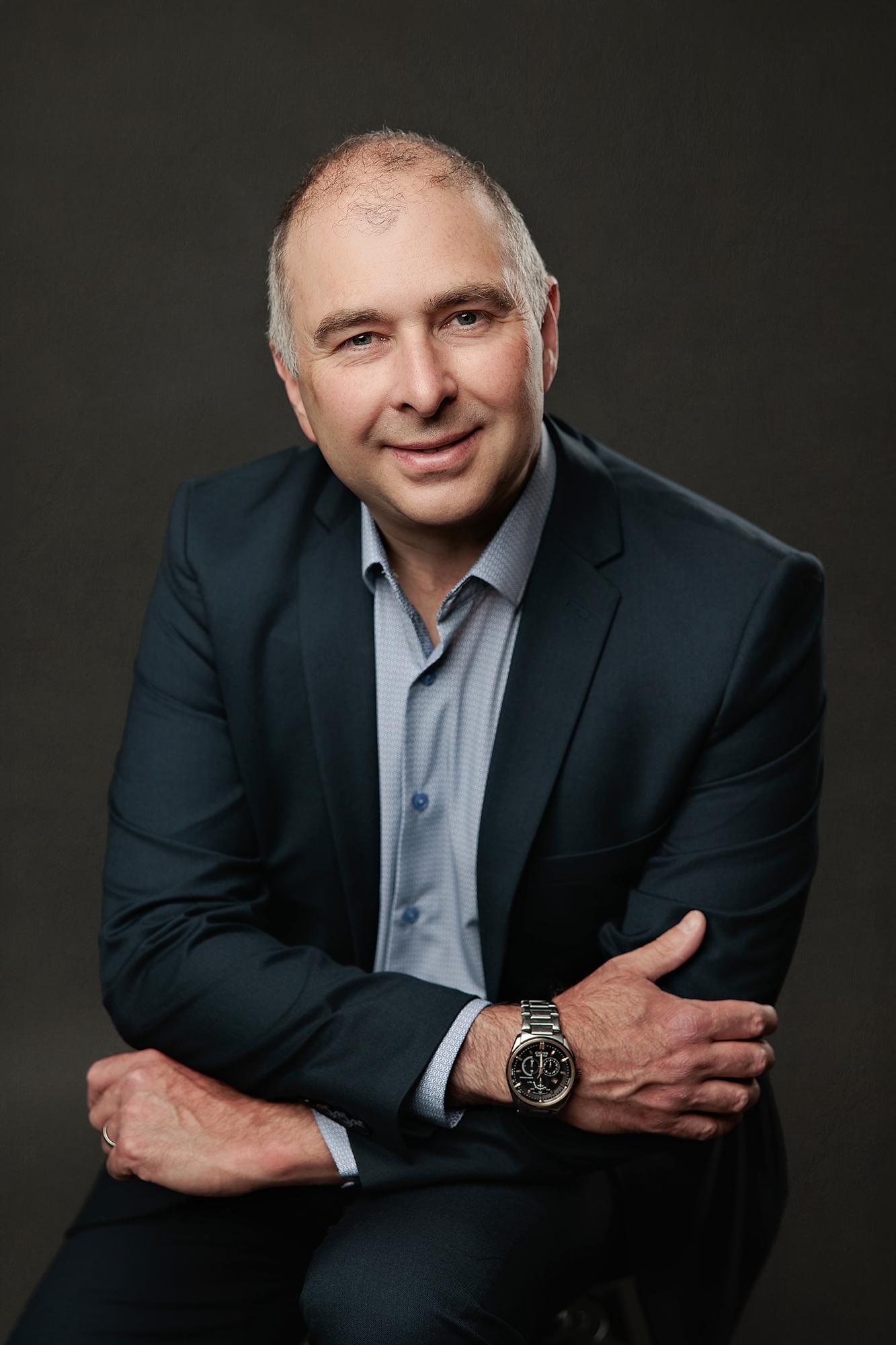 A smiling man in a navy blazer poses for a professional headshot at our Calgary studio, featuring soft studio lighting against a dark textured backdrop.