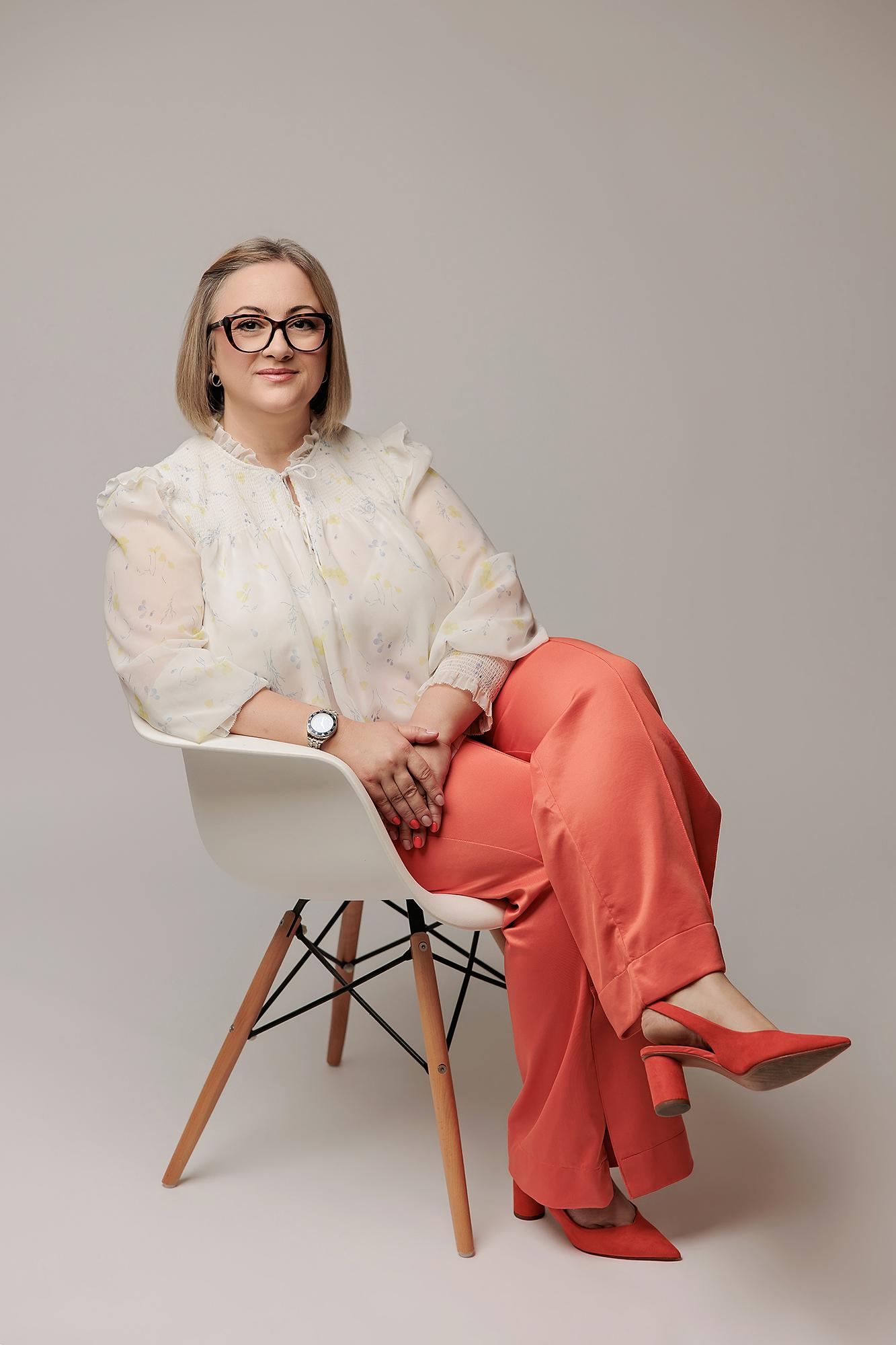 A professional woman in a floral blouse and coral trousers sits in a Calgary photography studio, exuding confidence for her personal branding session.