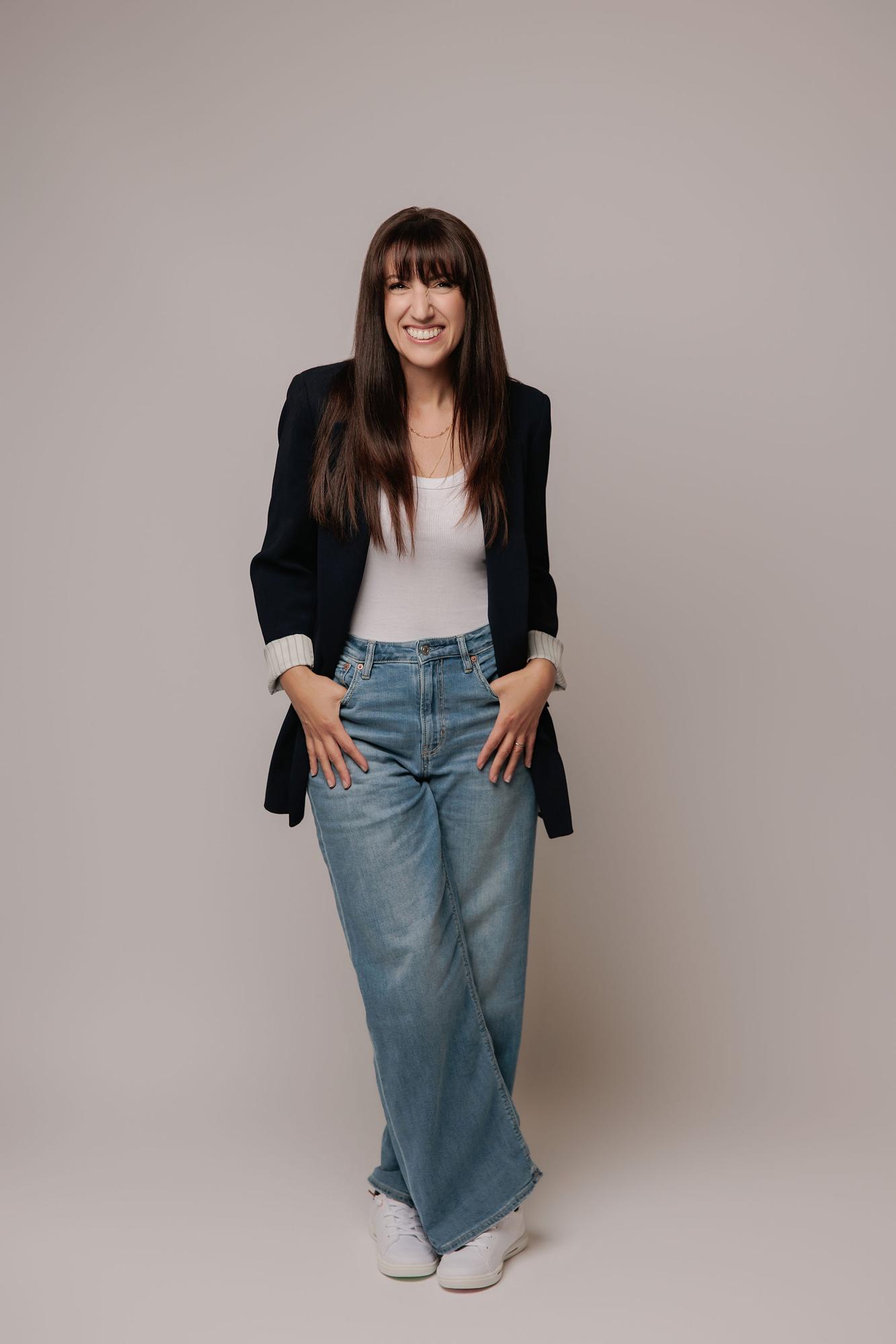 A woman with long dark hair smiles in a navy blazer and jeans, showcasing the relaxed professional style of a Calgary headshot photography studio.