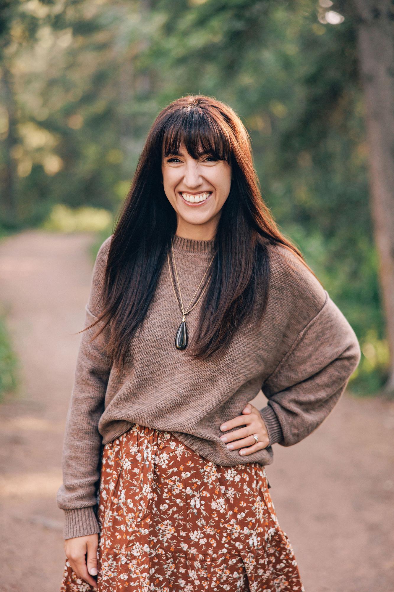 A smiling woman wears a brown sweater and floral skirt in a natural outdoor setting, captured by a Calgary photographer for professional personal branding.