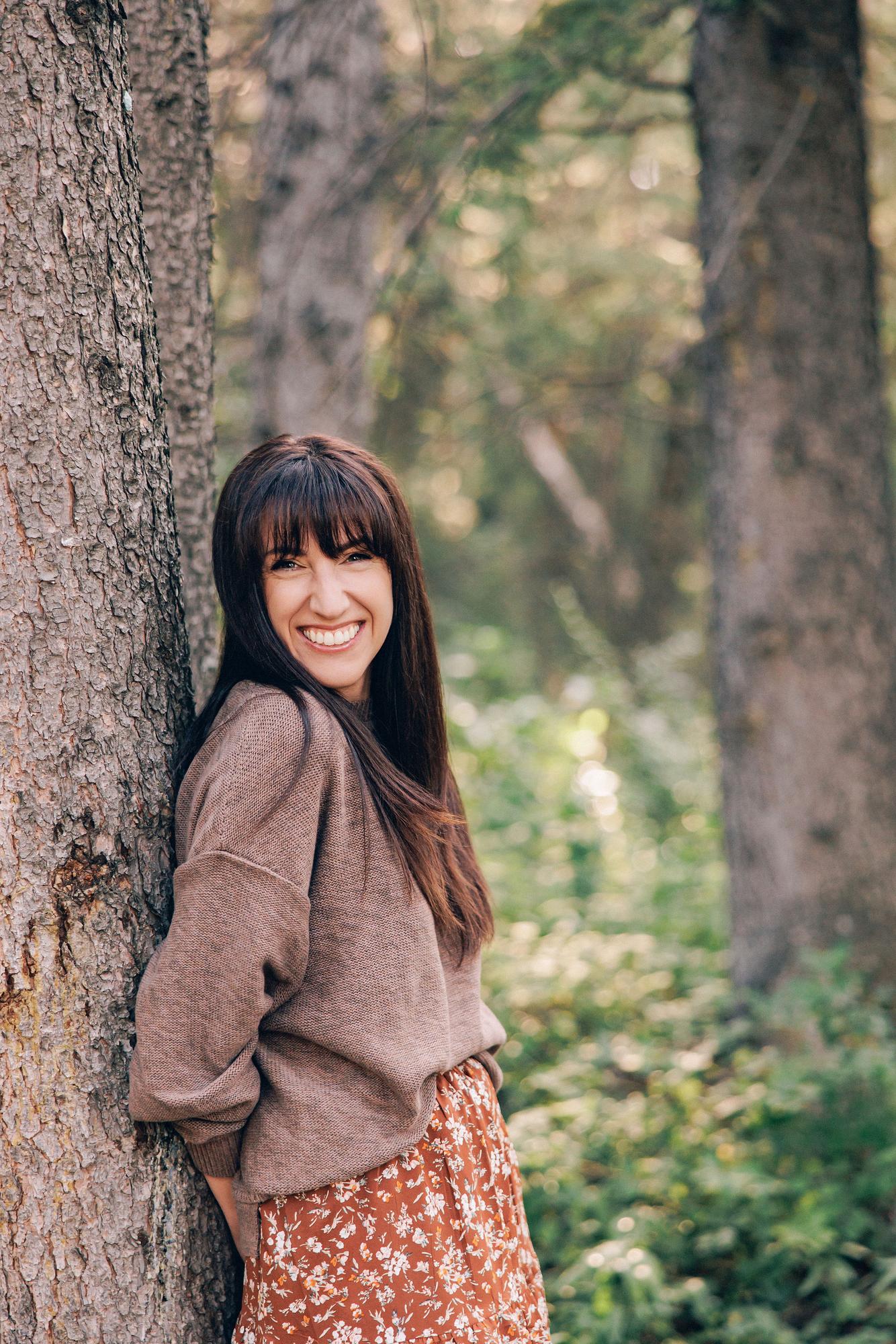 A smiling woman in a cozy brown sweater leans against a pine tree in a forest, showcasing the natural outdoor style of a Calgary headshot photographer.