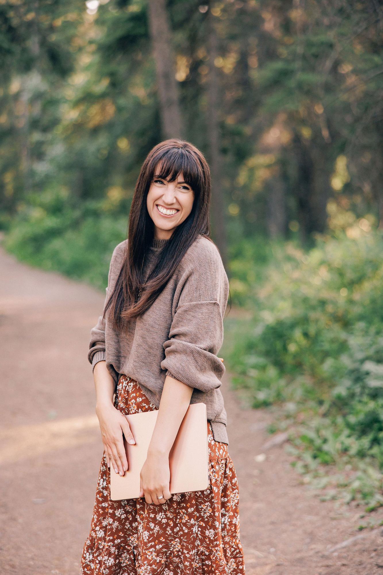 A woman with bangs smiles while holding a laptop in a lush park, showcasing the natural outdoor style of a professional Calgary personal branding photographer.