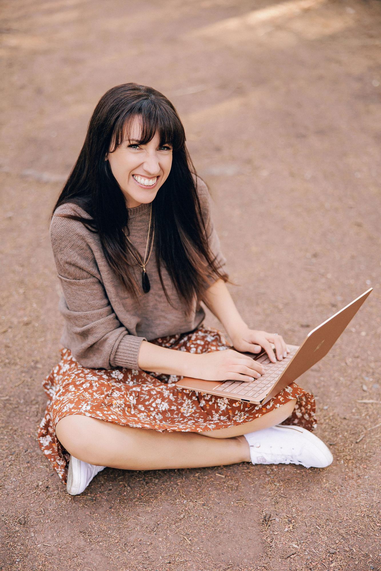 A smiling woman in a brown sweater and floral skirt works on a laptop outdoors, showcasing the natural style of this Calgary personal branding photographer.