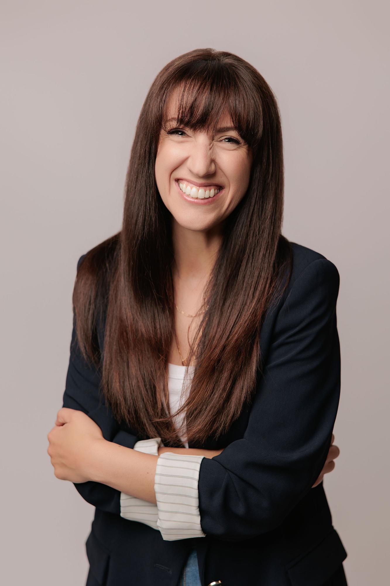 A professional woman with long brown hair smiles brightly in this Calgary headshot session, wearing a navy blazer against a clean studio backdrop.