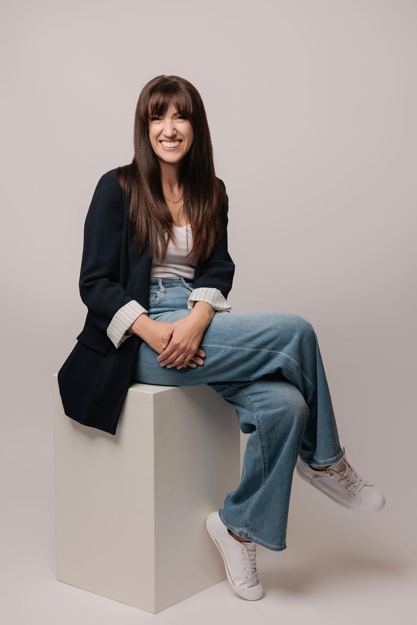 A woman with long brown hair smiles while sitting on a white cube, showcasing the relaxed personal branding style of our Calgary photography studio.