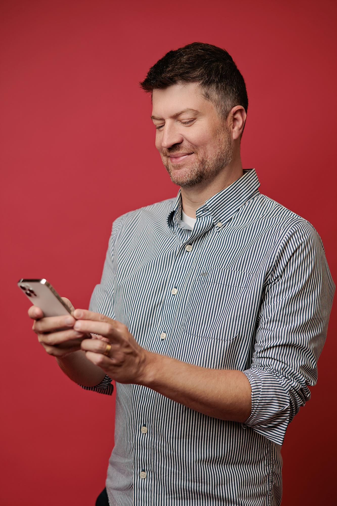 A man in a striped button-down shirt smiles while using his phone in this Calgary photography studio portrait, captured against a vibrant red backdrop.