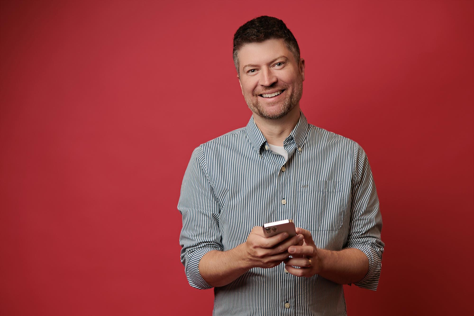 A smiling man in a striped button-down shirt holds a smartphone against a vibrant red backdrop, captured by a Calgary headshot photographer.