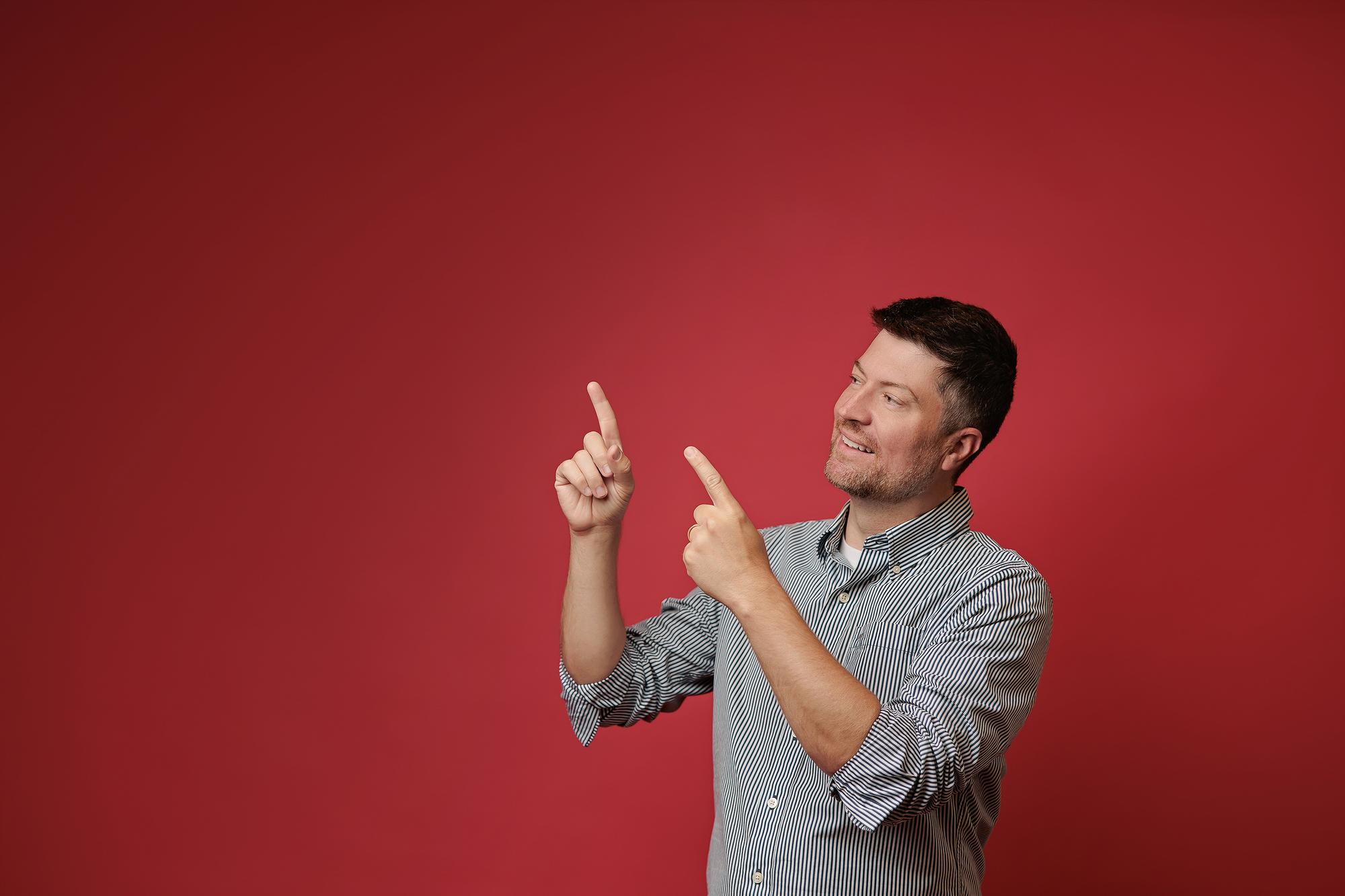 A man in a striped button-down shirt smiles while pointing upward, captured in a Calgary photography studio with a vibrant red background.