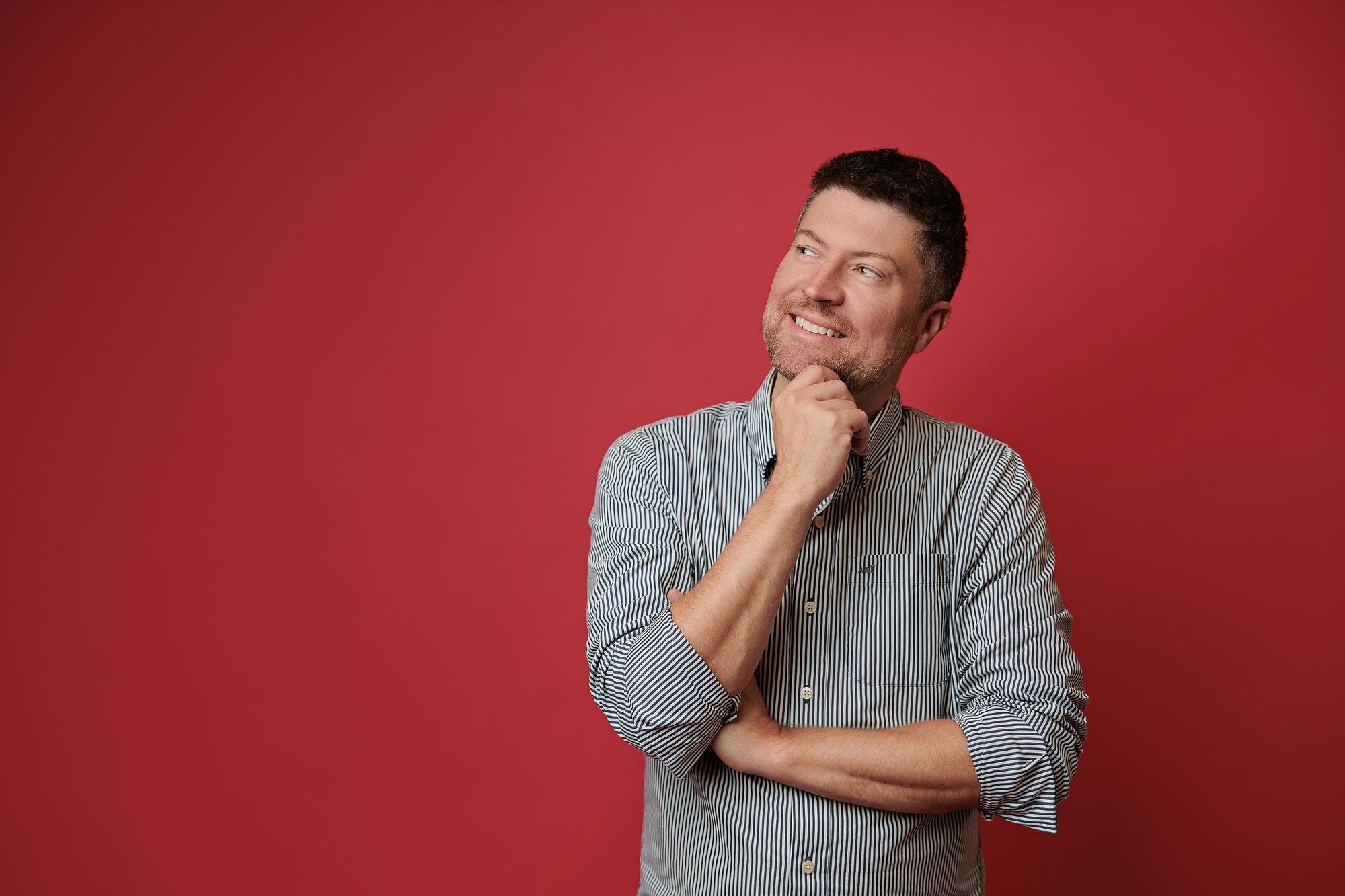 A man in a striped shirt poses thoughtfully against a vibrant red backdrop, showcasing the creative portrait style of this Calgary headshot photographer.