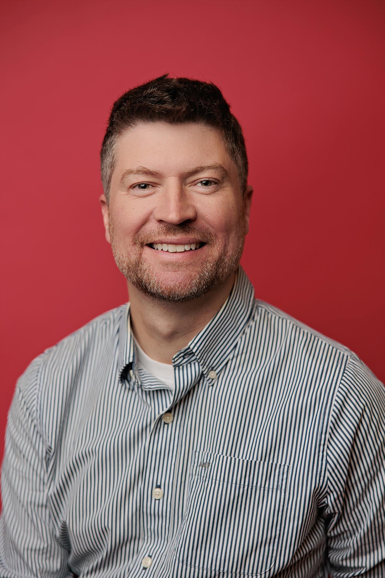 A smiling man in a striped button-down shirt poses against a vibrant red backdrop, showcasing the bold style of a professional Calgary headshot studio.