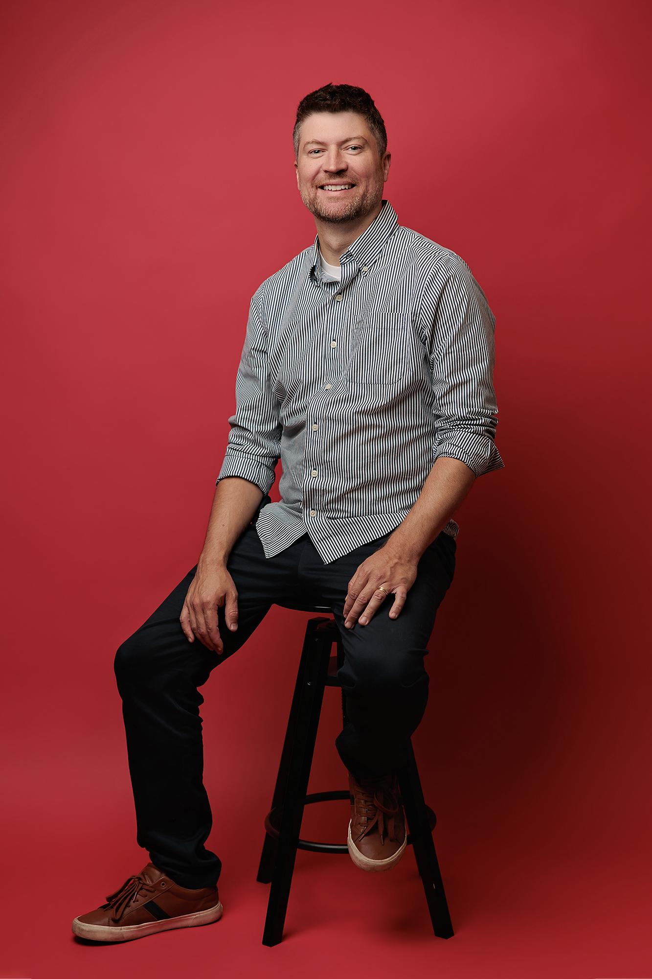 A smiling man in a striped button-down shirt sits on a stool against a vibrant red backdrop, showcasing the bold style of our Calgary photography studio.