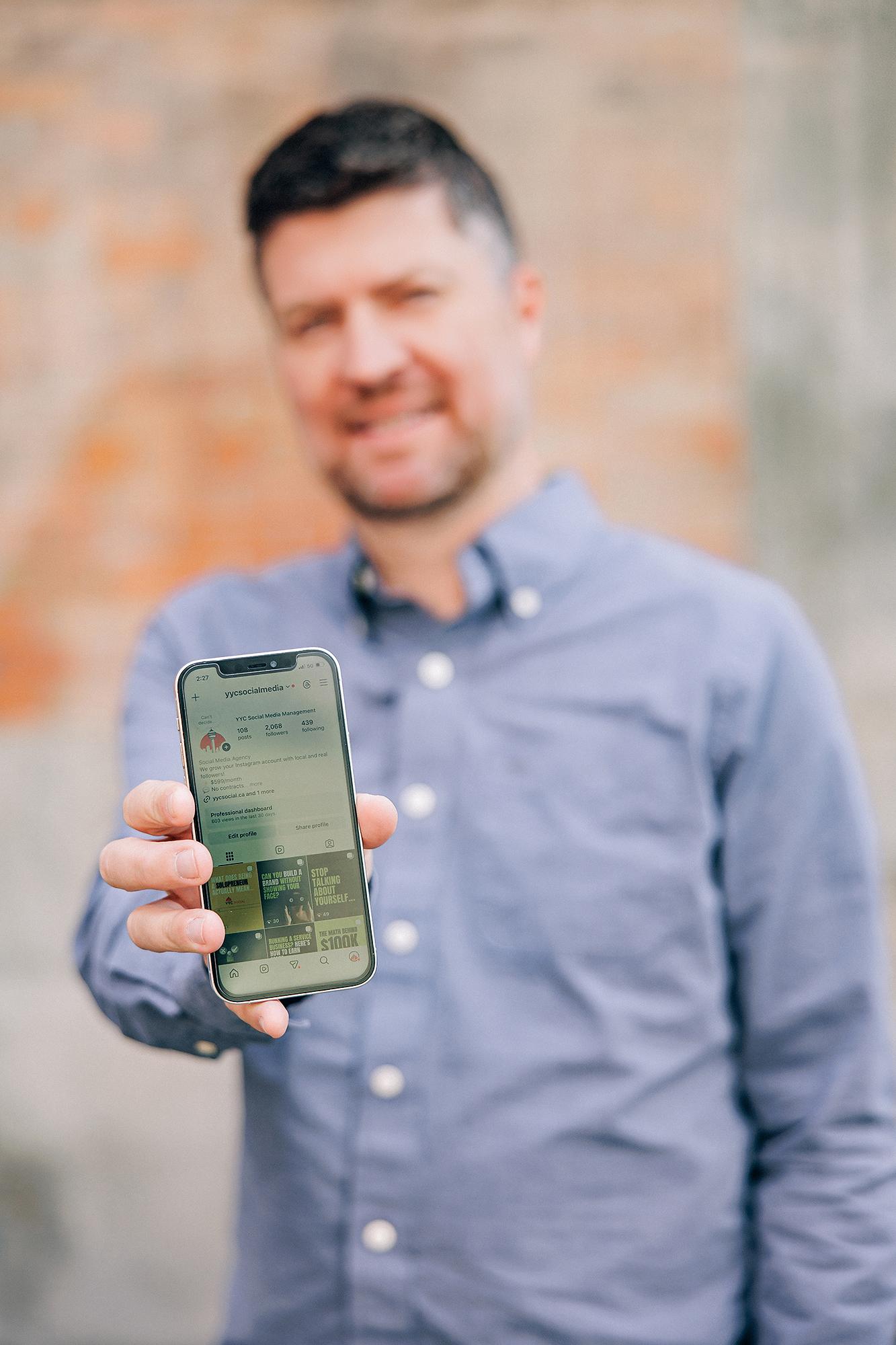 A smiling man in a blue button-down shirt holds a phone displaying a social media profile, showcasing personal branding photography by our Calgary studio.
