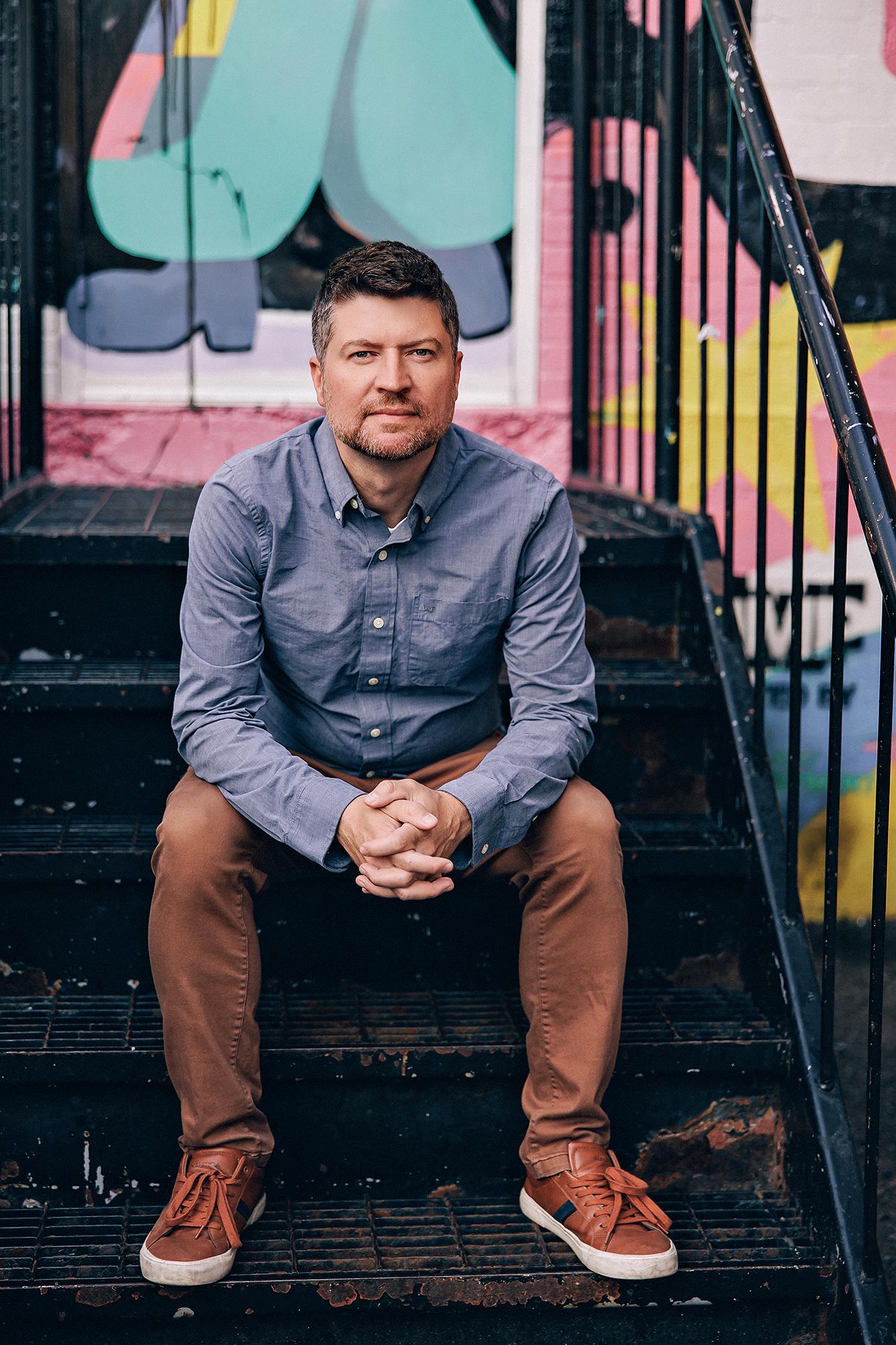 A man in a blue button-down shirt sits on metal stairs for a Calgary branding session, showcasing the creative style of our local photography studio.
