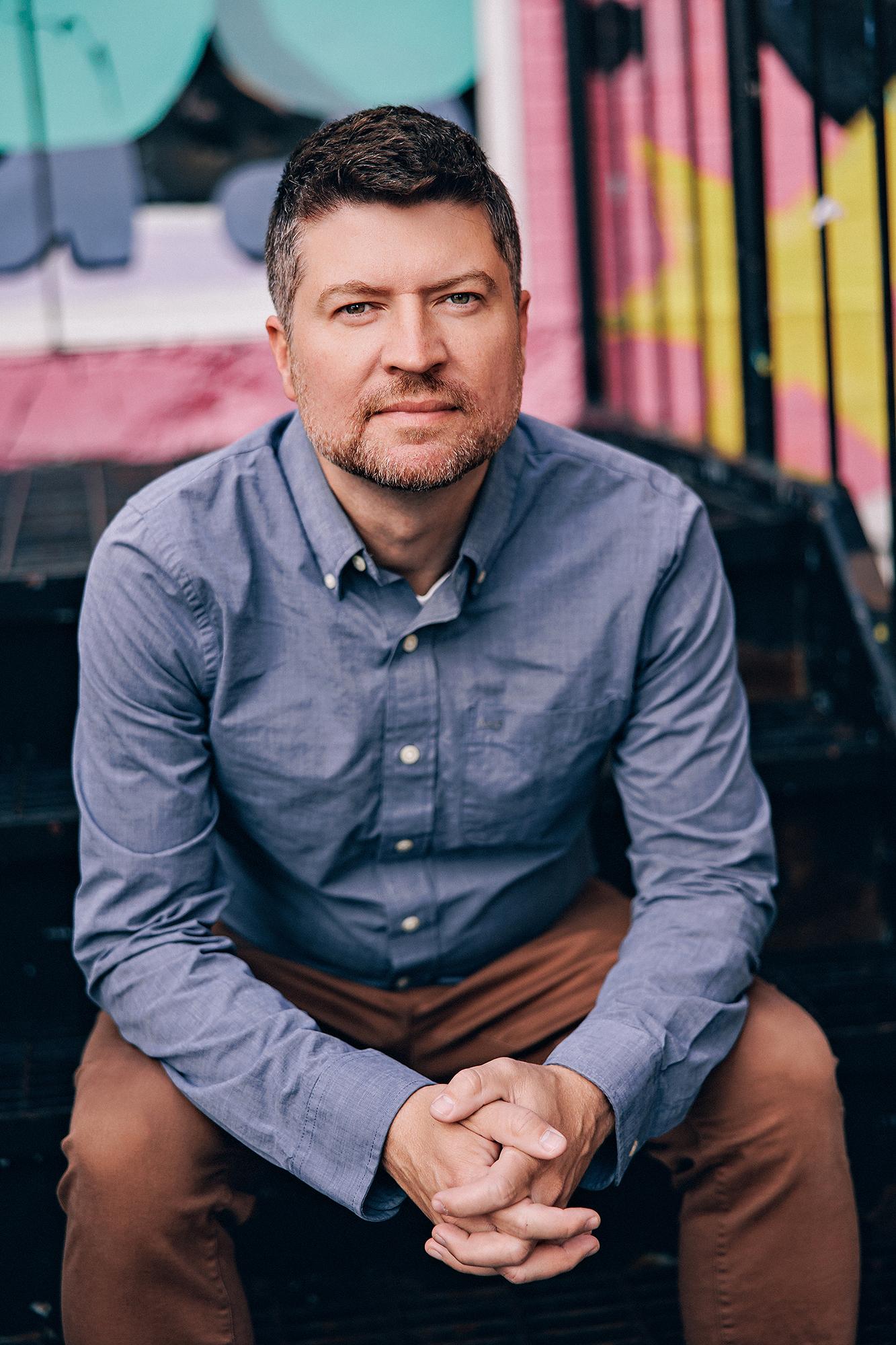 A man with a calm expression wears a blue button-down shirt while sitting on outdoor stairs, captured by a Calgary headshot photographer for branding.
