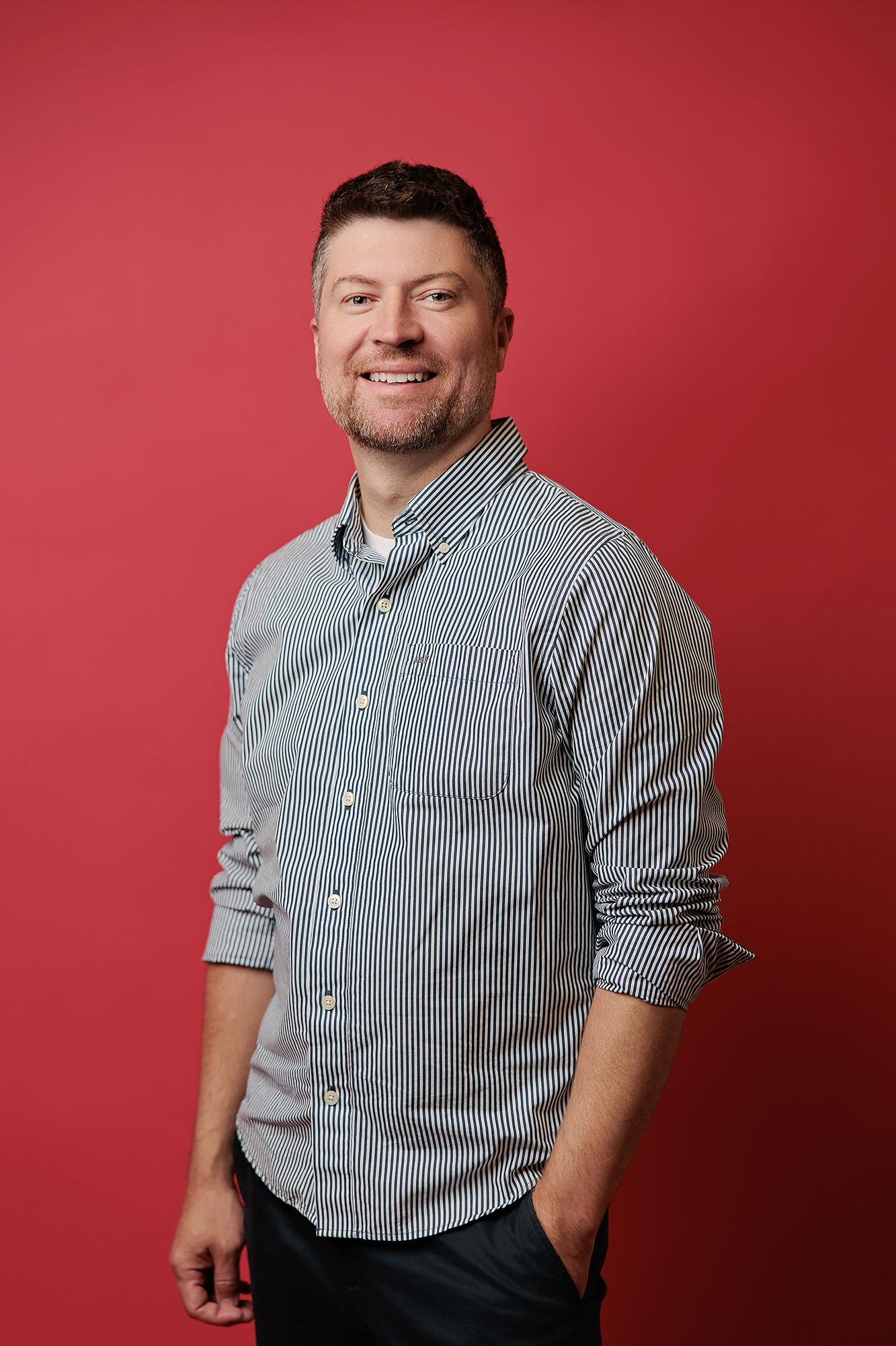A smiling man wears a striped button-down shirt against a vibrant red backdrop in this professional headshot from our Calgary photography studio.