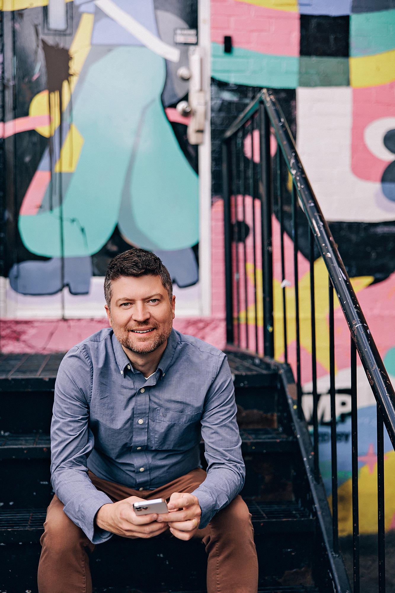 A smiling man in a blue button-down shirt sits on metal stairs for a Calgary personal branding session, captured by a professional studio photographer.