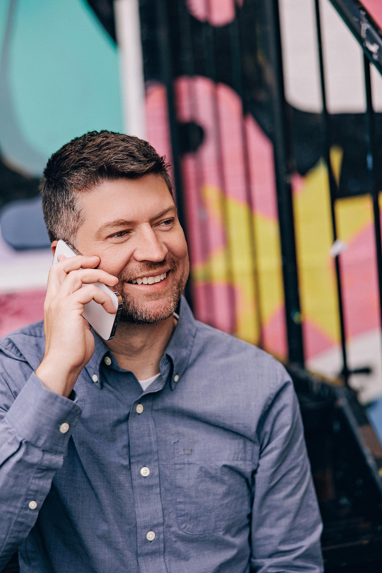 A smiling man in a blue button-down shirt talks on his phone, captured by a Calgary headshot photographer against a vibrant, urban graffiti backdrop.