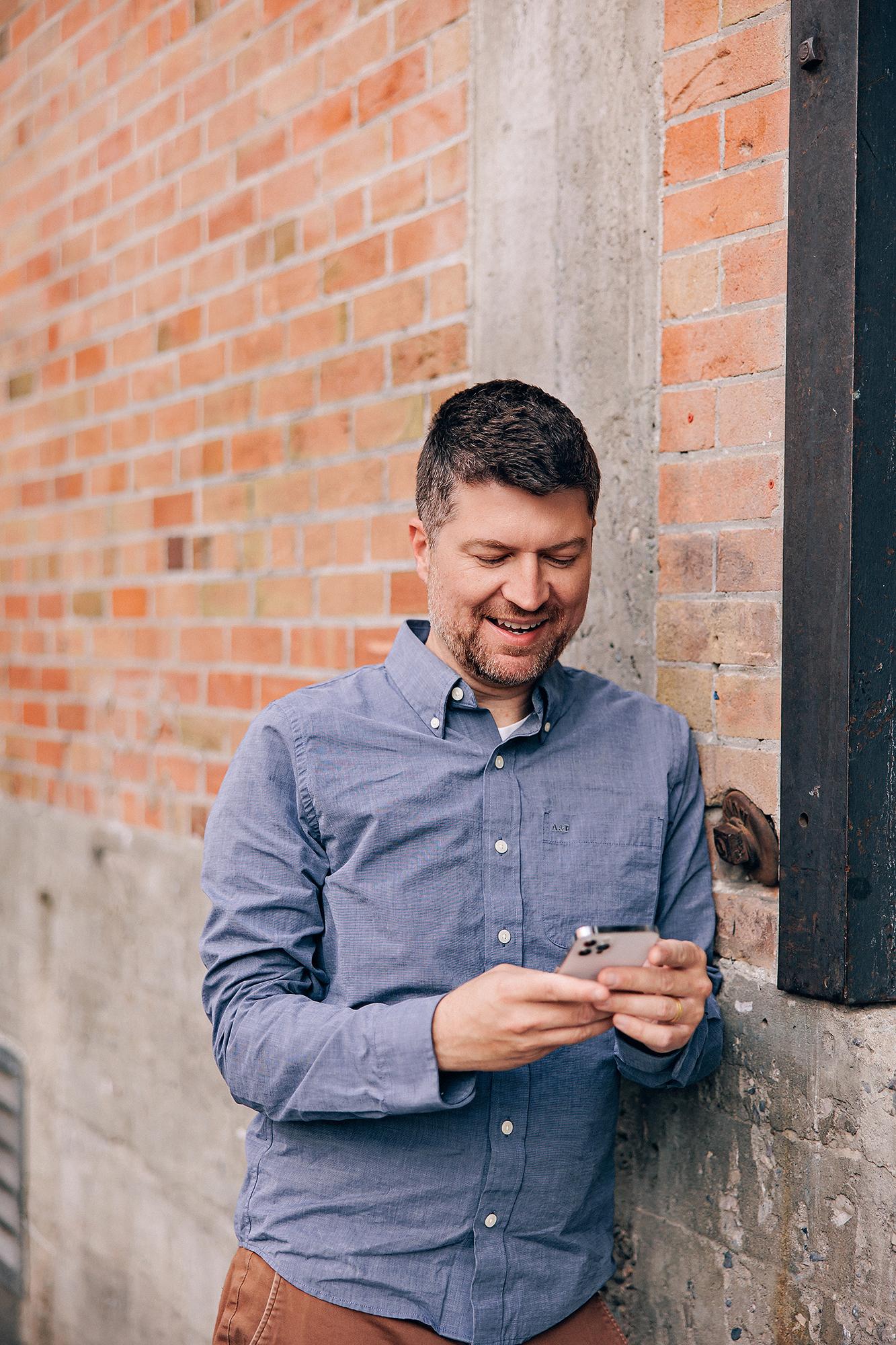 A man in a blue button-down shirt smiles while looking at his phone, captured by a Calgary headshot photographer against a rustic brick wall.